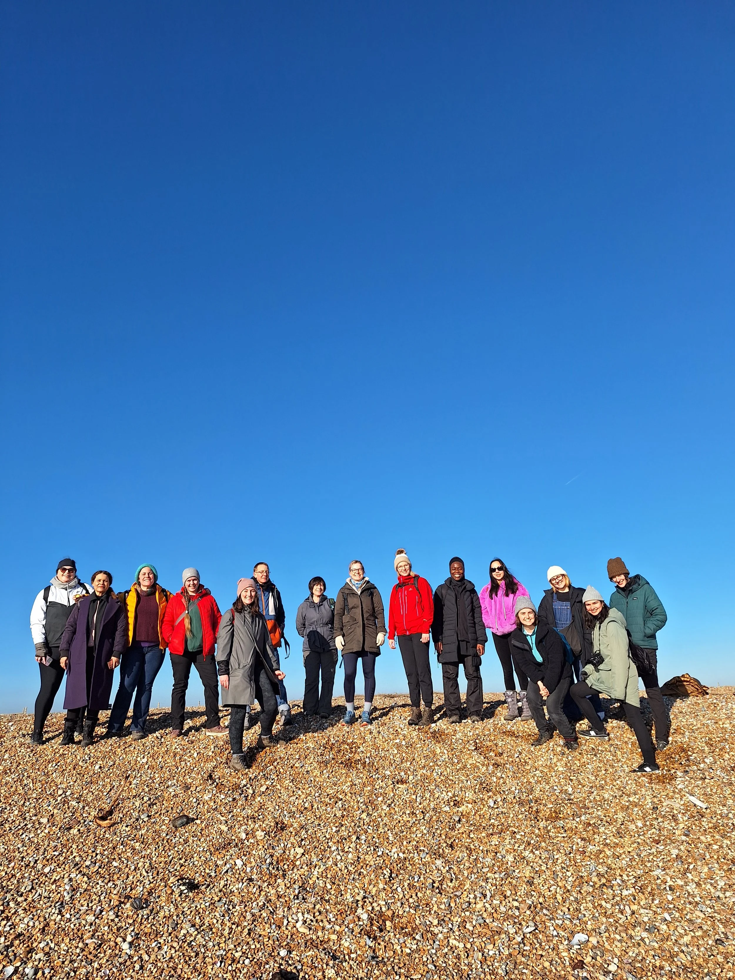 A group of fifteen women standing together on a shingle beach under a clear blue sky, dressed in outdoor hiking or casual winter clothing, smiling for a photo.