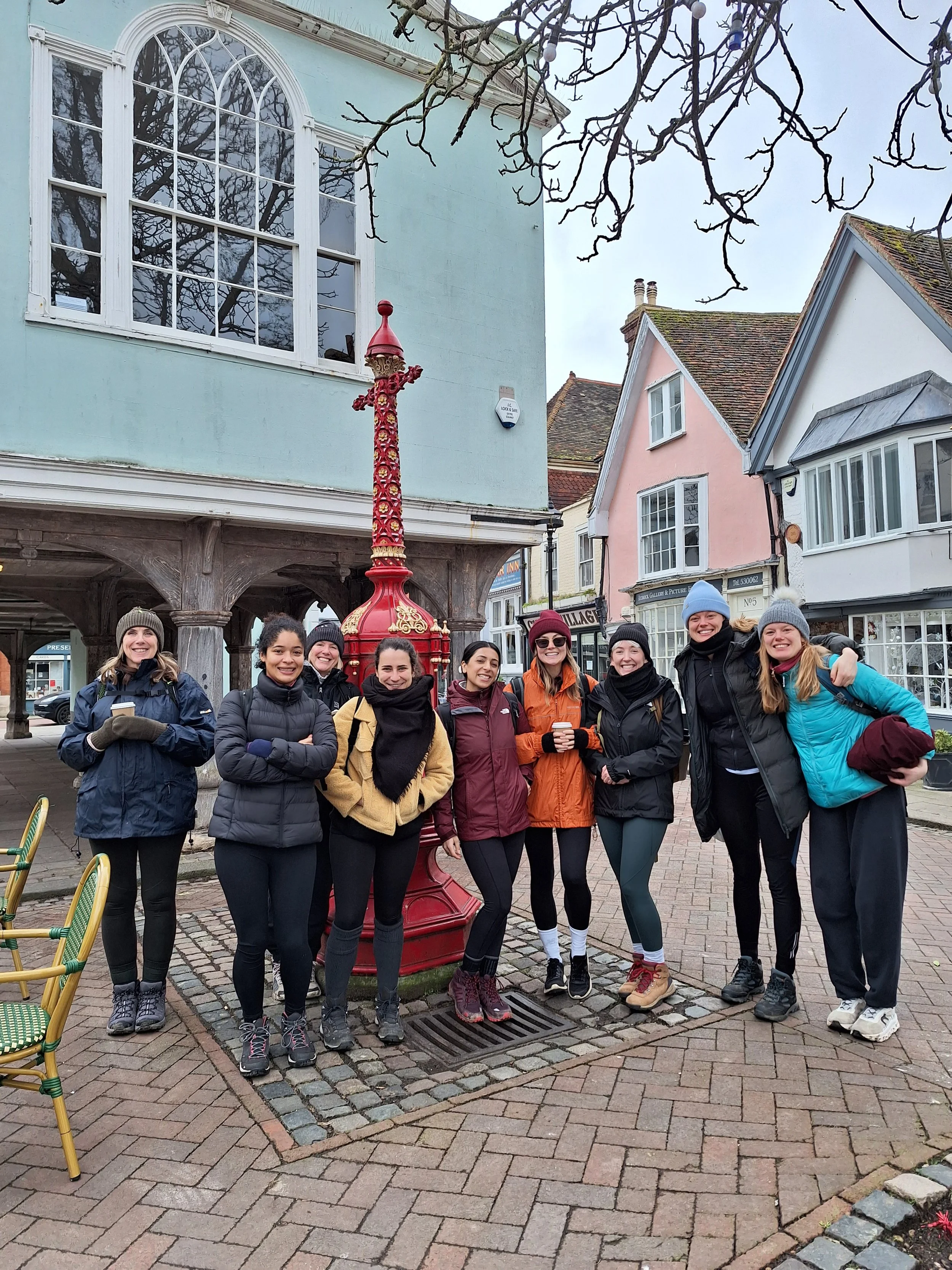 A group of ten women standing together outdoors in front of a historic-looking building, smiling and dressed in casual outdoor clothing, with a red ornate street lamp behind them.