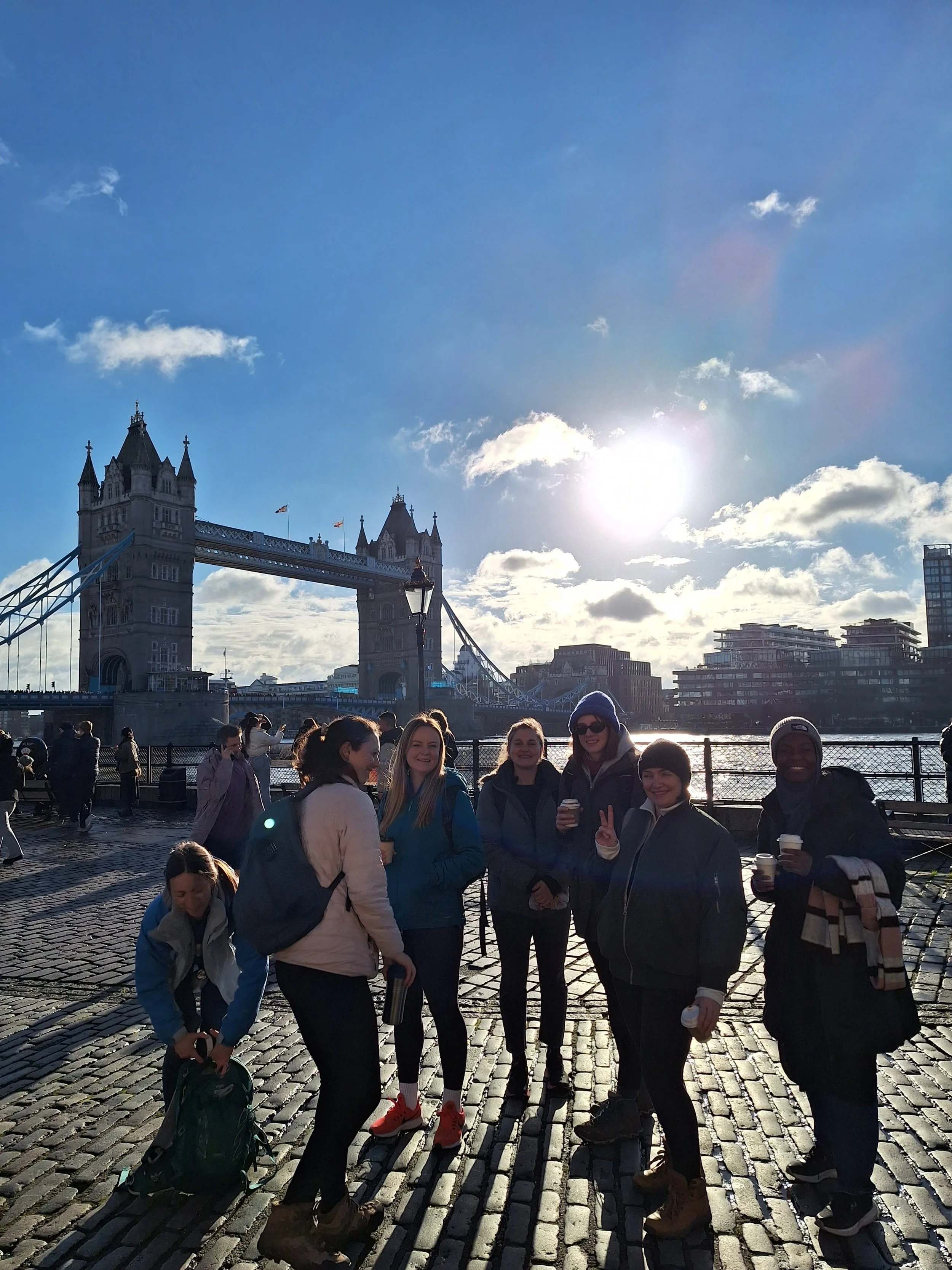 A group of women standing on a cobblestone bridge with Tower Bridge visible in the background during daytime, with sunlight and a partly cloudy sky.