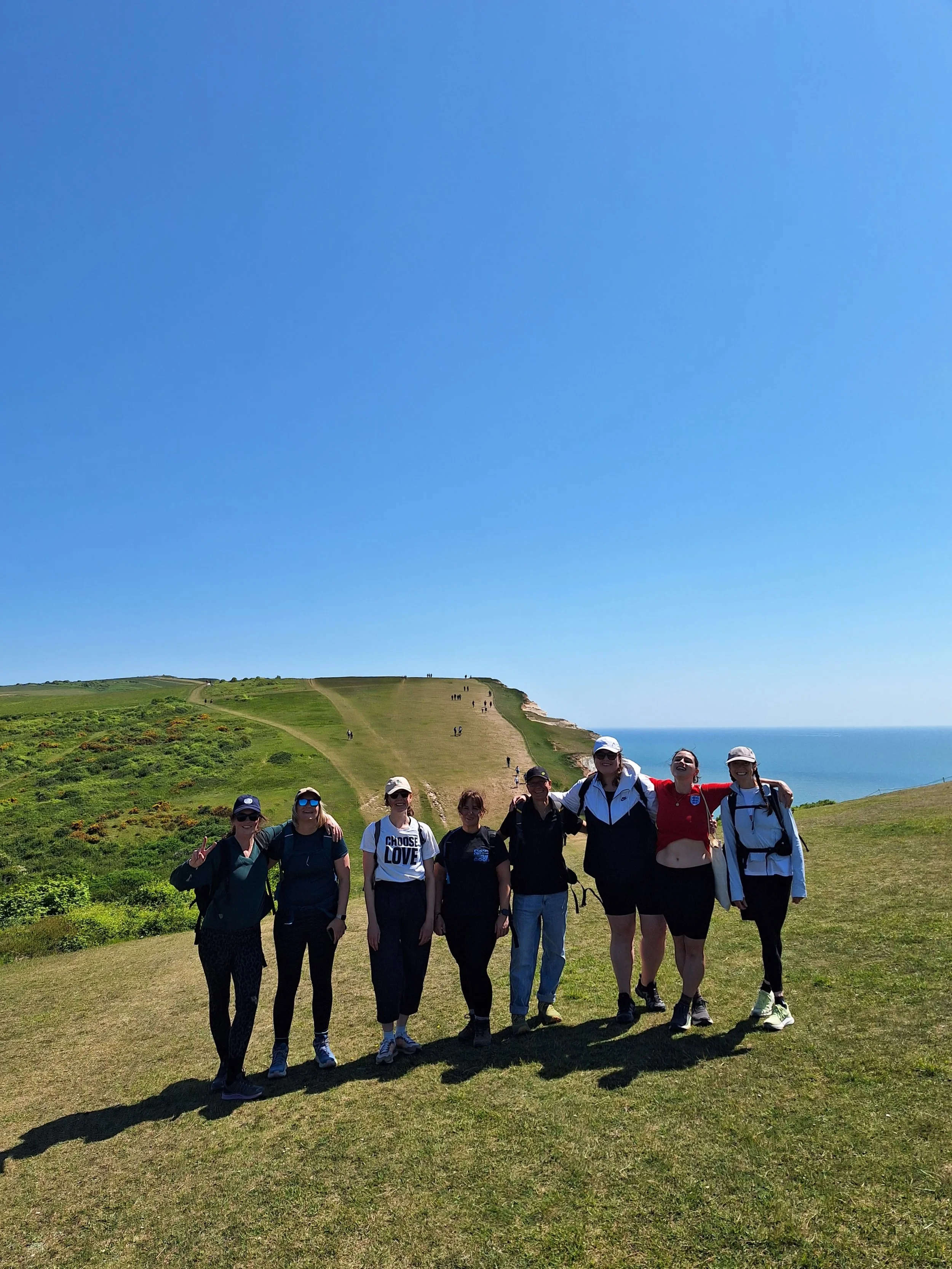 Group of women standing on a grassy hill with green cliffs and the sea in the background, under a blue sky.