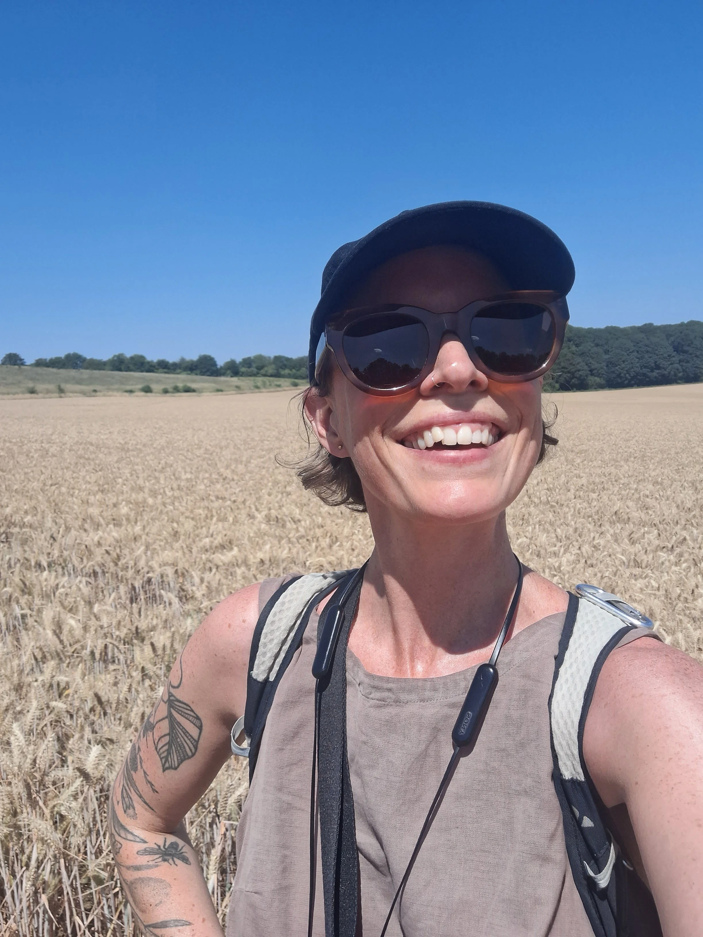 A smiling woman in a cap and sunglasses, standing in a golden crop field on a sunny day with a bright blue sky