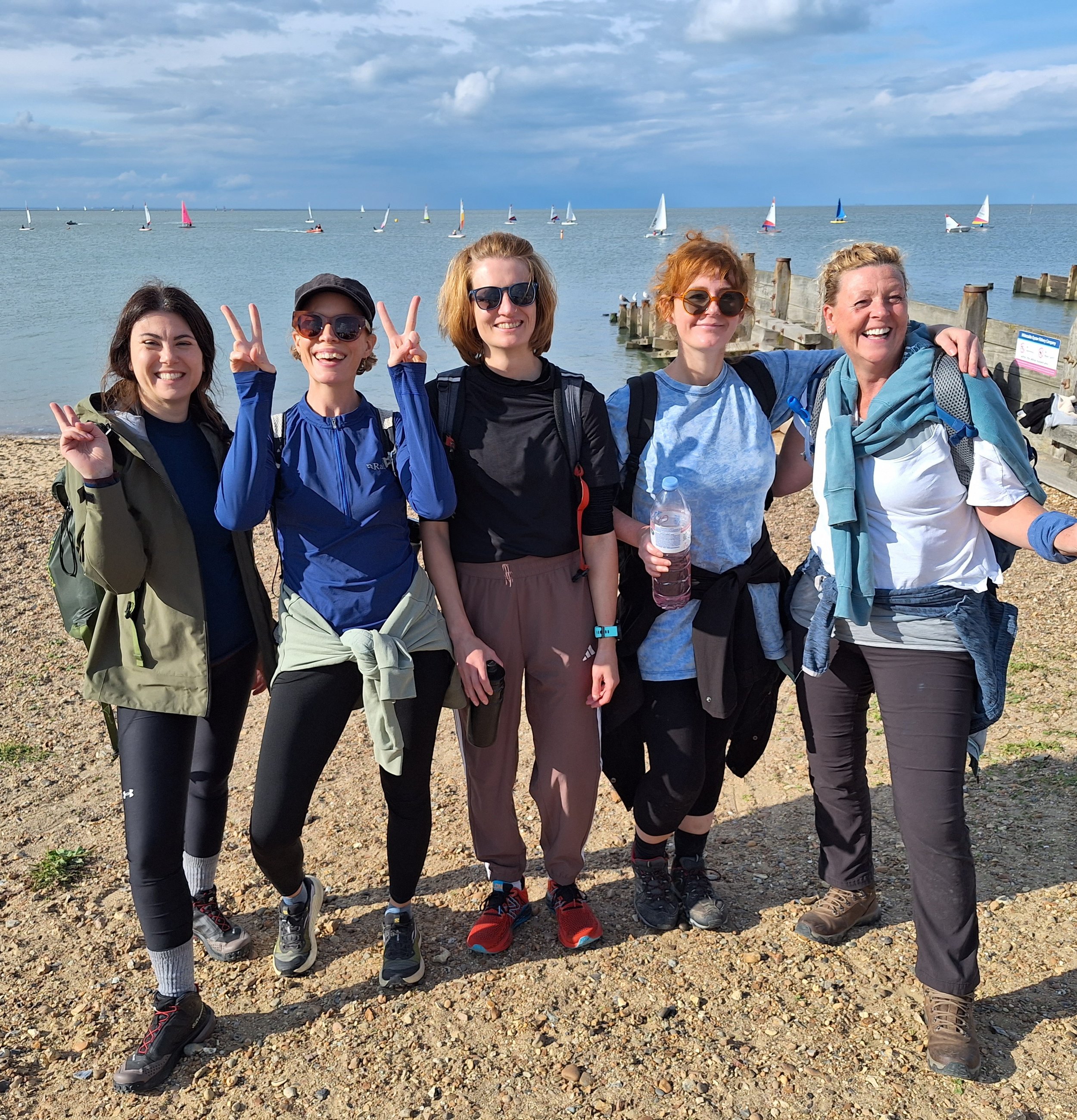 A group of five women standing on a rocky beach near a body of water with sailboats sailing in the background on a partly cloudy day. They are smiling, wearing outdoor clothing and backpacks, and some are making peace signs.