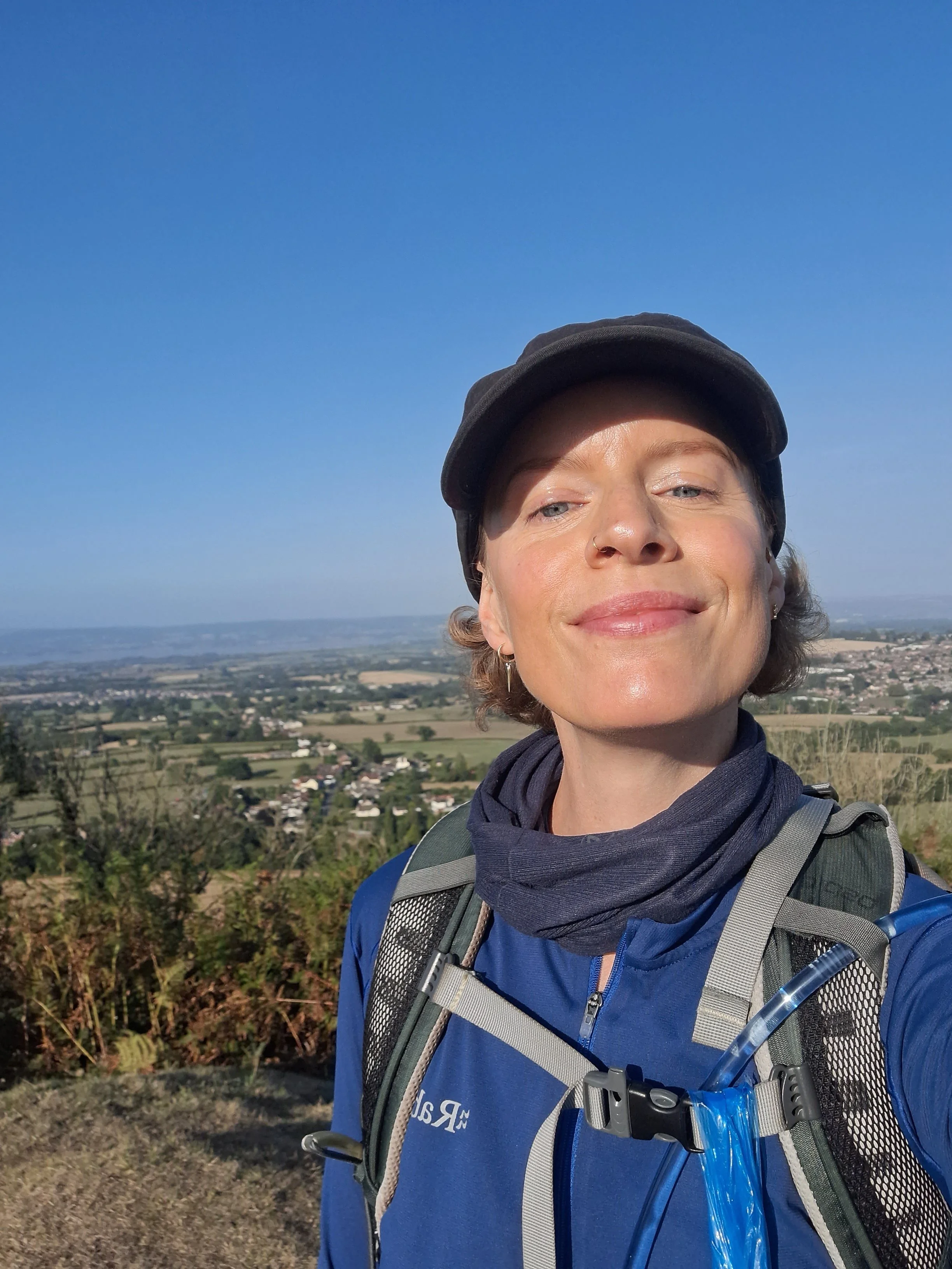 A smiling woman in sportswear and a cap, standing on a hill in the sunshine, with a sweeping view of the countryside behind her