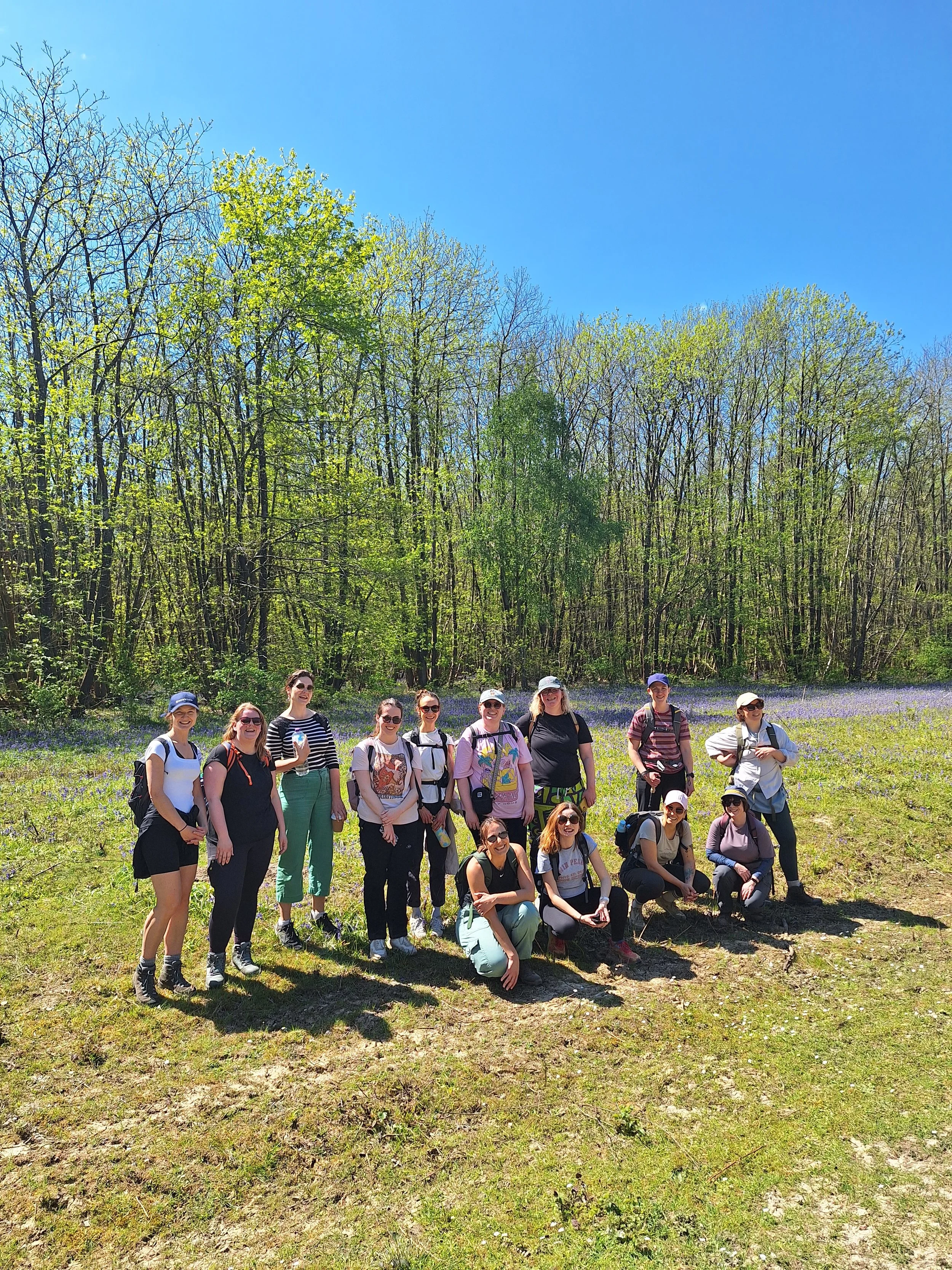 A group of about fifteen women standing outdoors on a grassy area with trees, bluebells and a blue sky in the background. They are smiling and dressed casually, some wearing backpacks, hats, and sunglasses.