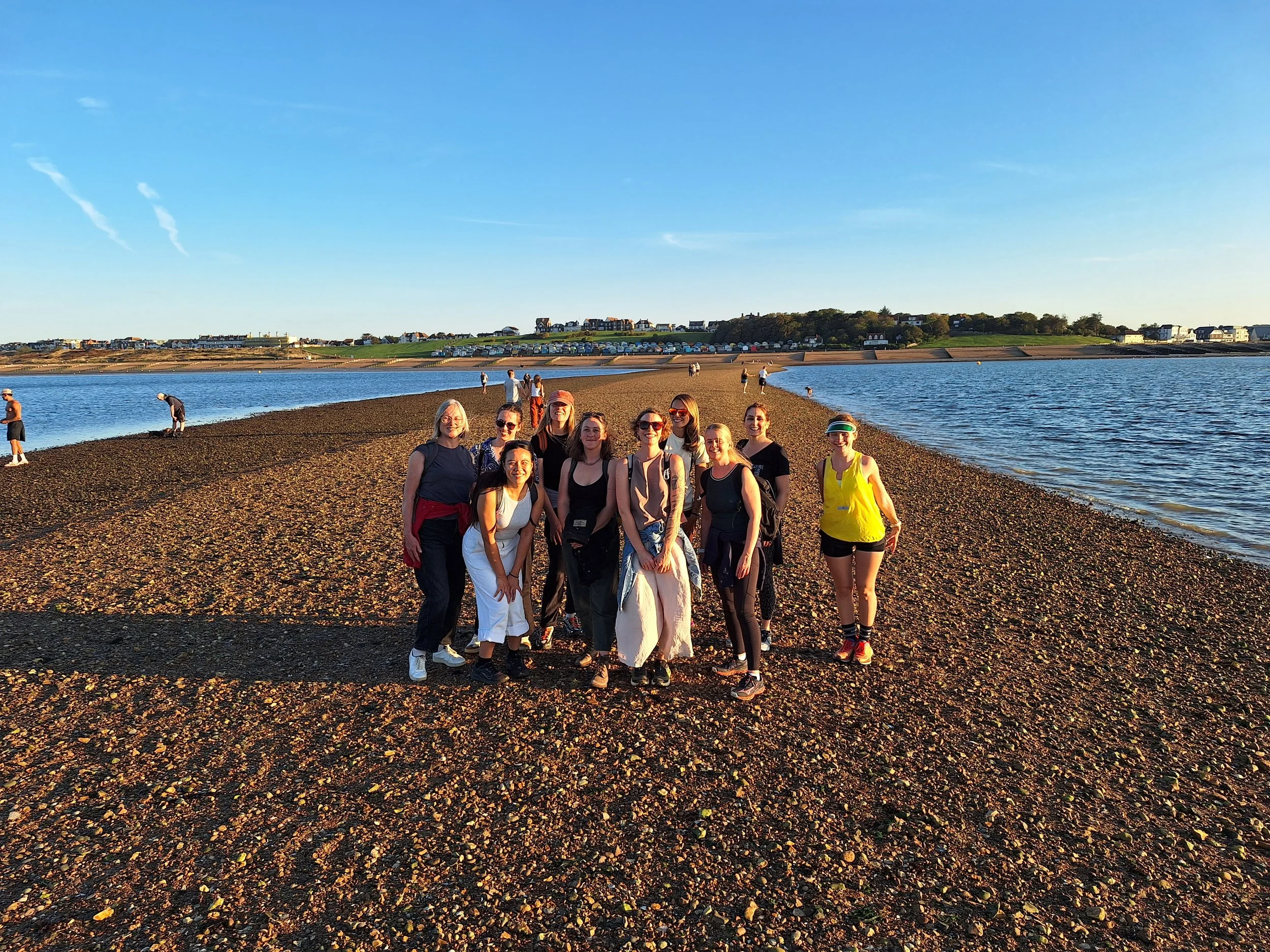 A group of women standing on a pebble beach near a body of water with houses on a hill in the background, during sunset.