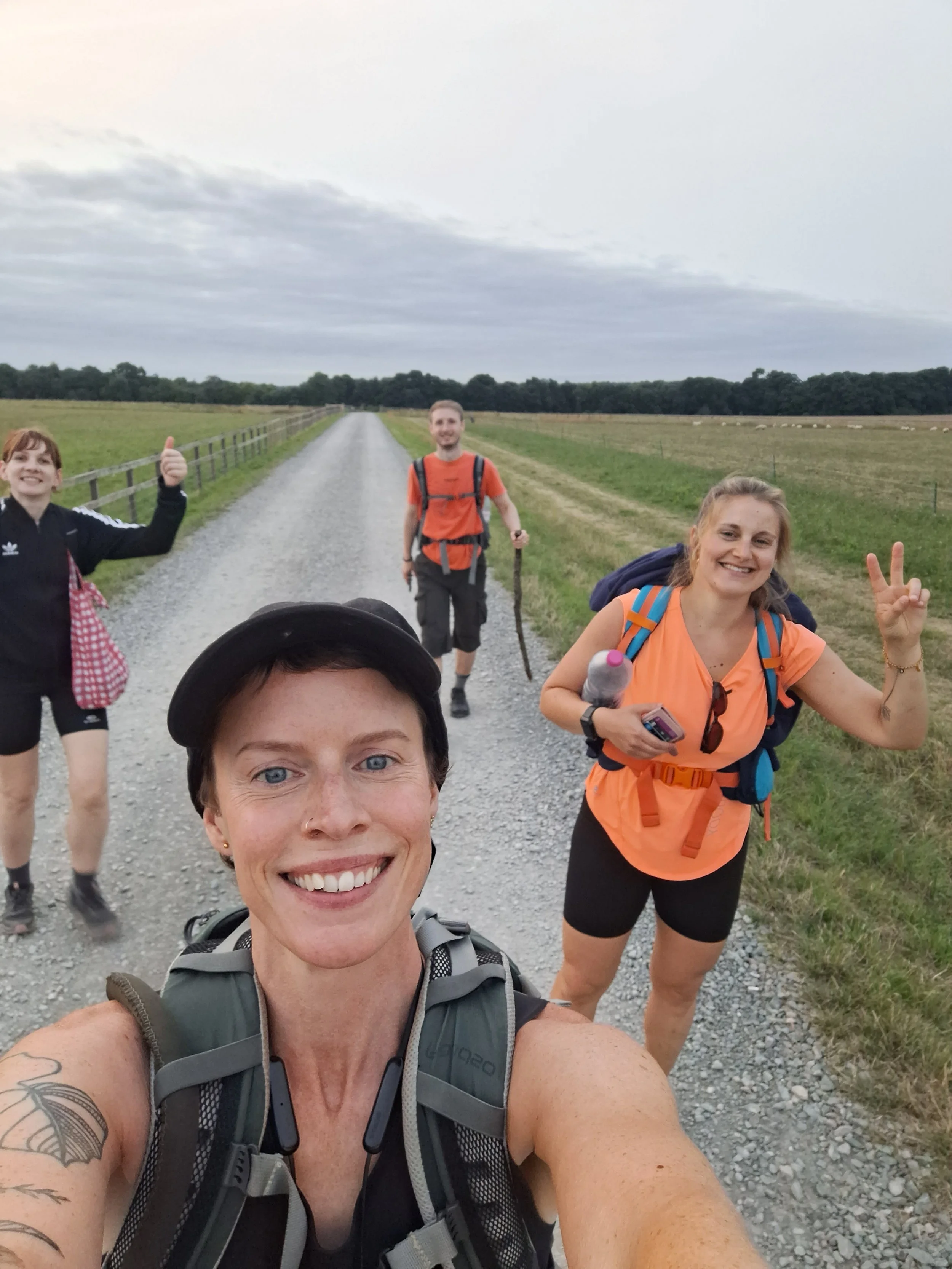 A group of four hikers walking on a gravel path through a rural area with fields and trees in the background, smiling and posing for a selfie.