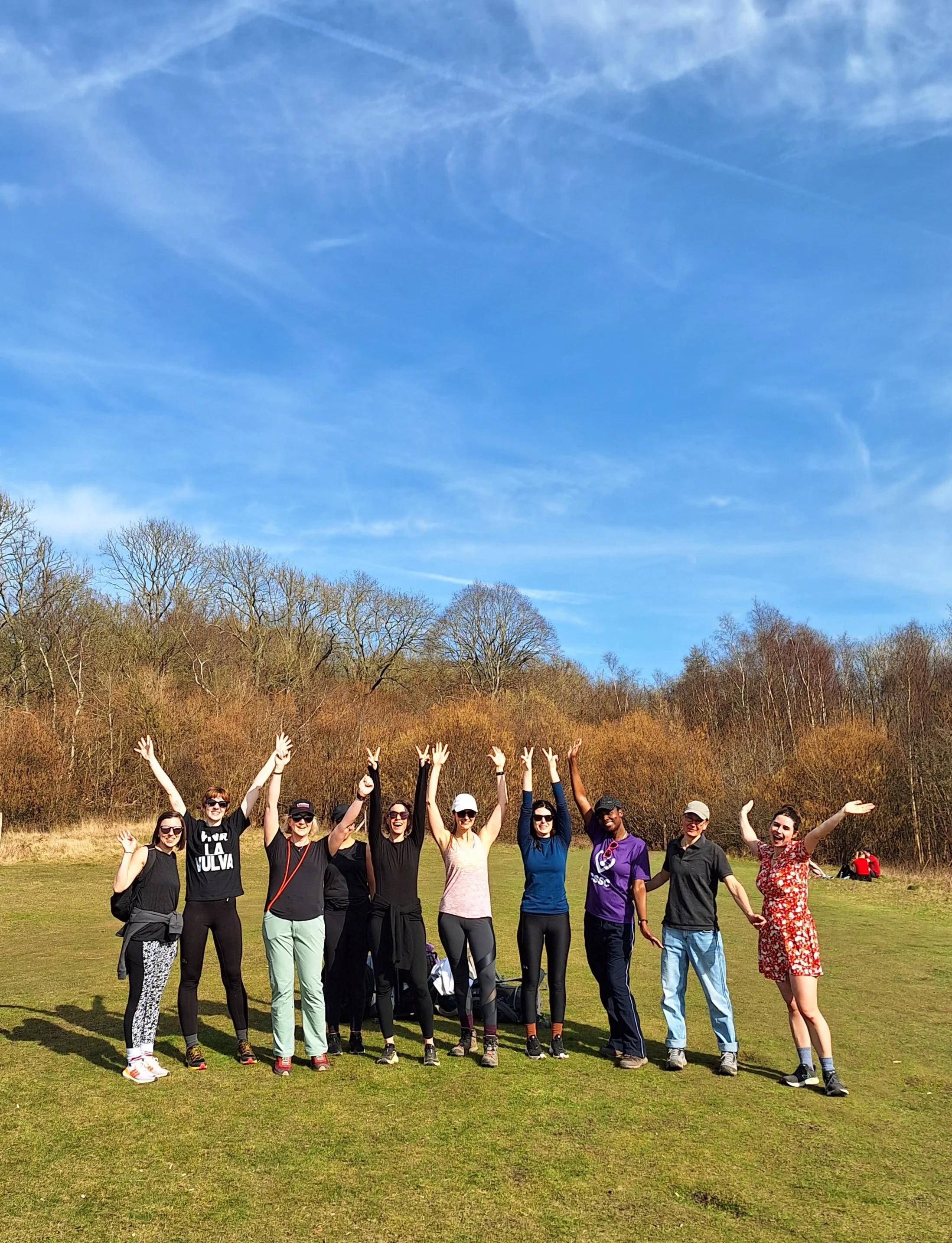 A group of women standing outdoors on grass, raising their hands and smiling, with leafless trees and blue sky in the background.