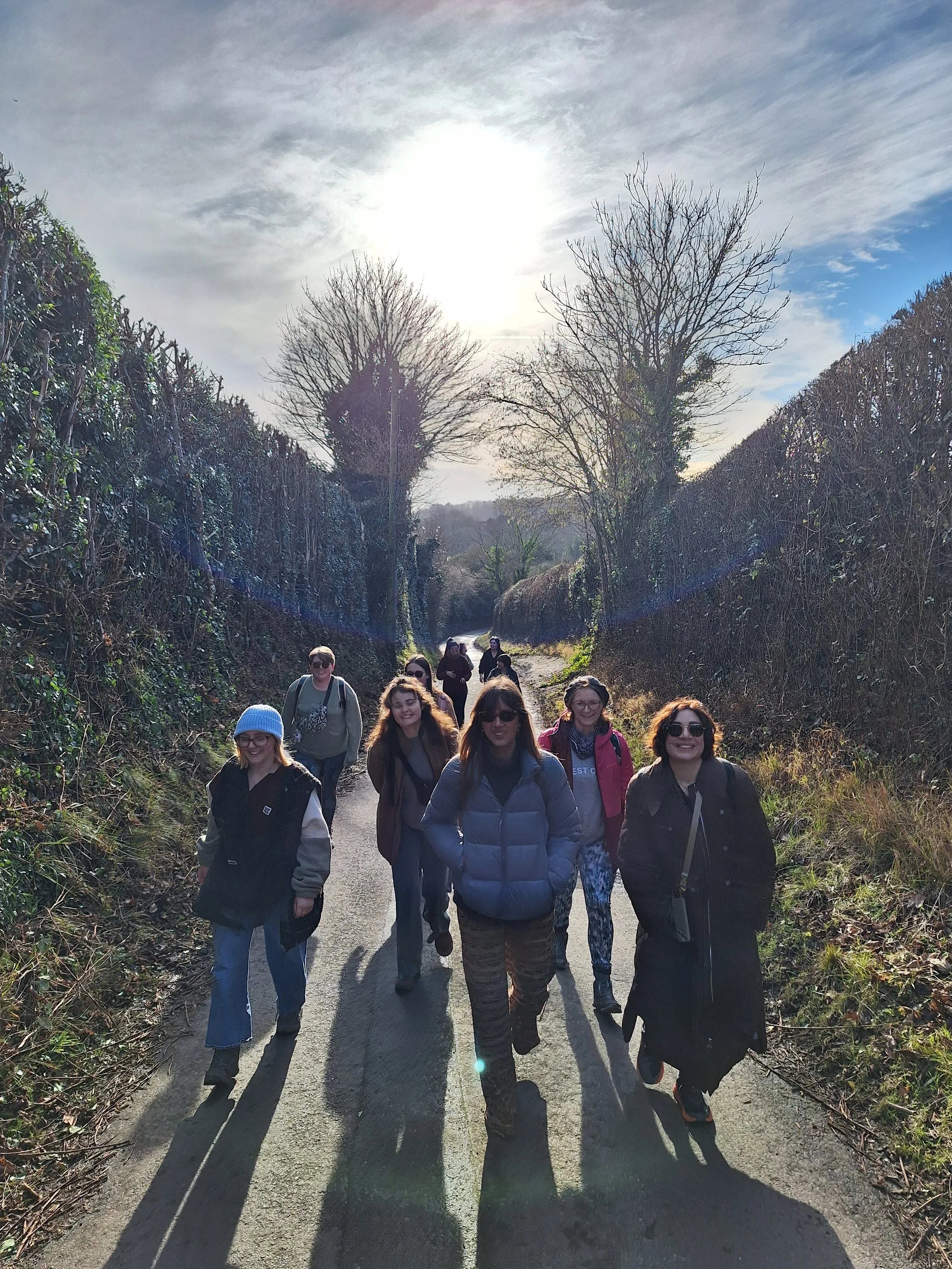 A group of women walking on a rural path surrounded by greenery and trees with bare branches. The sun is shining brightly in a partly cloudy sky, casting long shadows.
