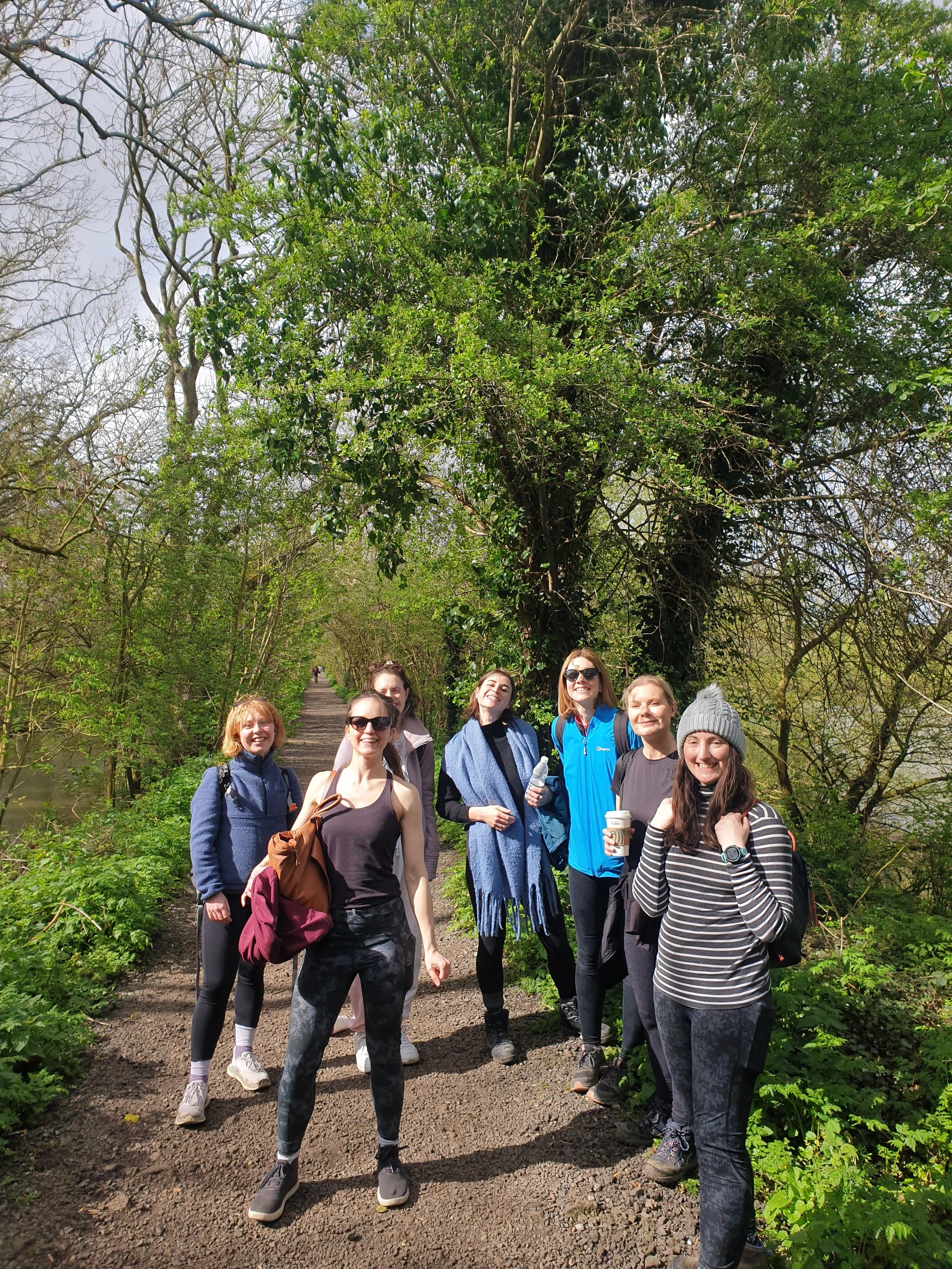A group of seven women standing on a dirt path in a lush green forest, smiling and posing for a photo. They are dressed in casual hiking clothes and carrying backpacks, with trees and foliage surrounding them.