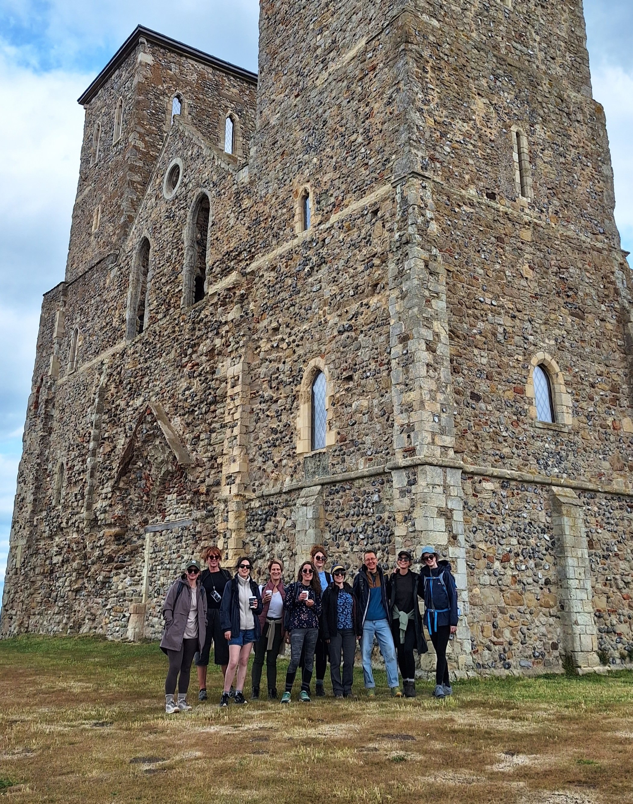 A group of women posing in front of a large stone tower with small windows, under a partly cloudy sky.