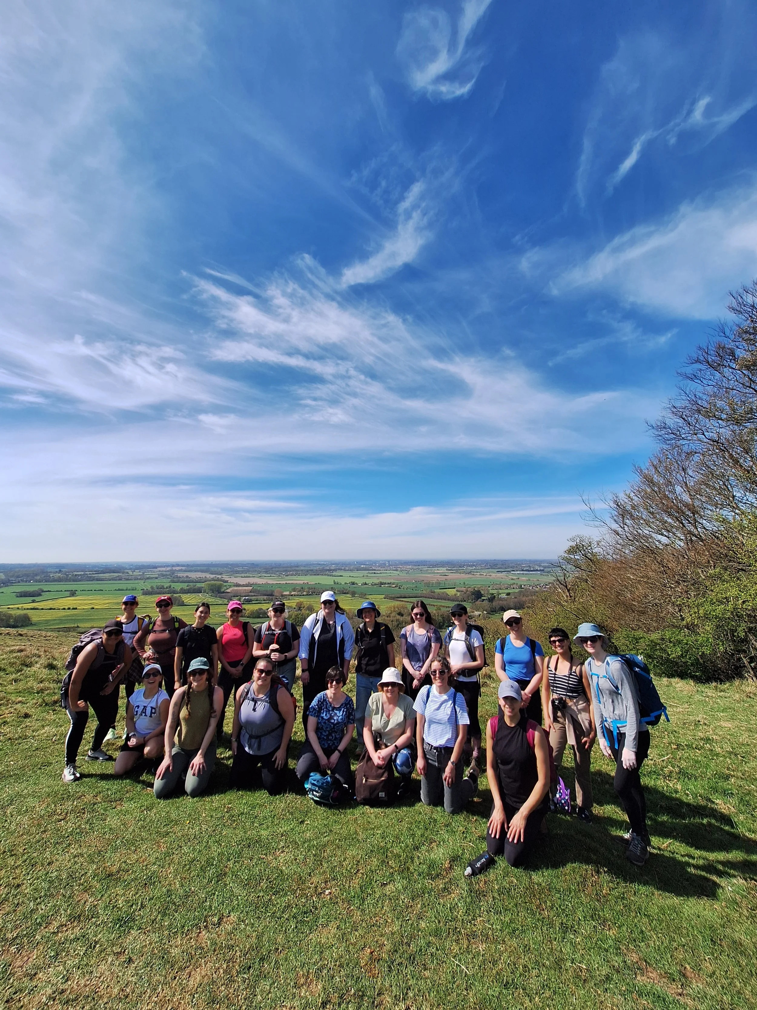 Group of women standing outdoors on a grassy hill with a scenic landscape and blue sky with clouds in the background.