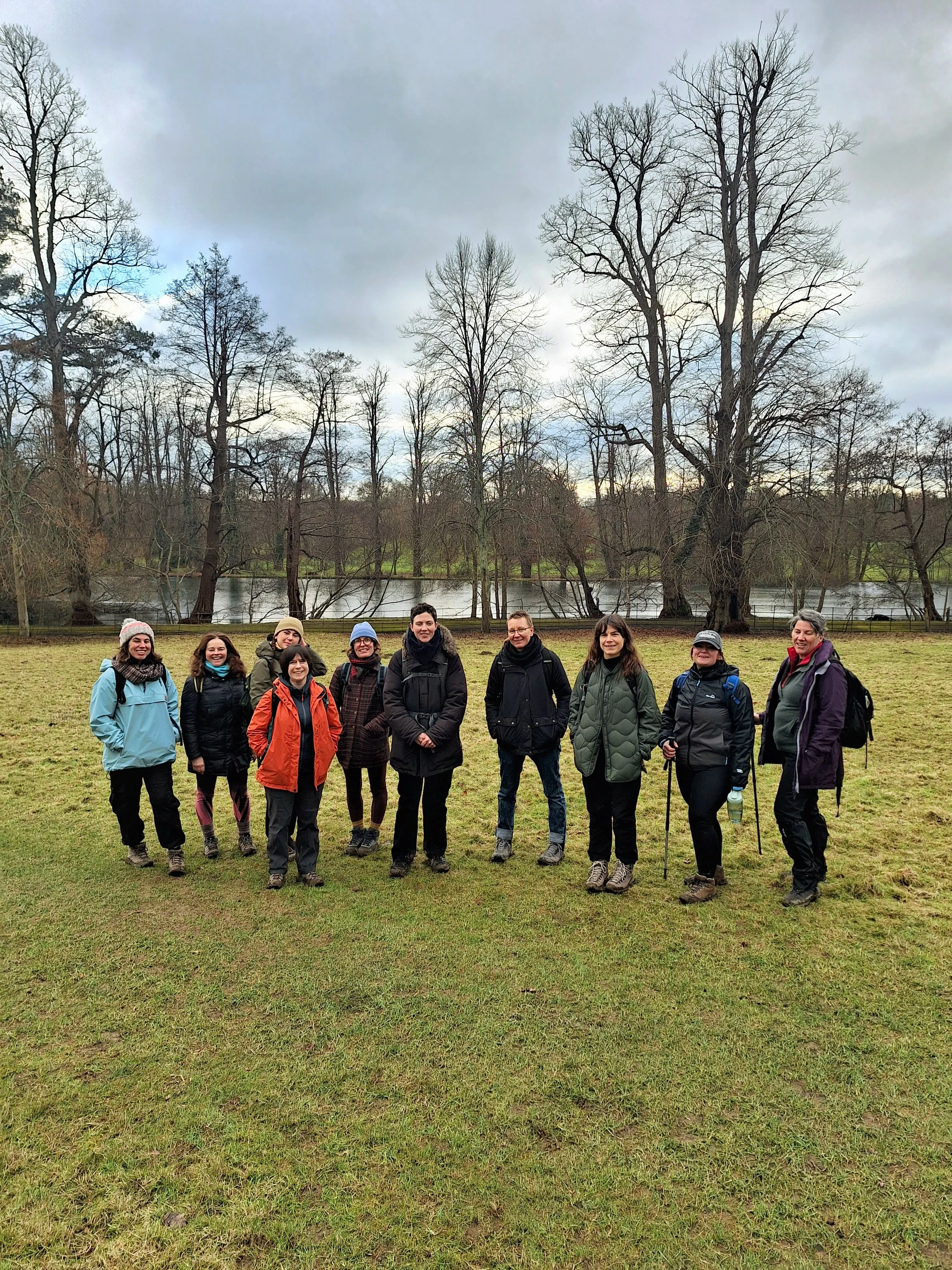 A group of eleven women standing outdoors on grass near a river, with leafless trees and cloudy sky in the background, dressed in outdoor clothing suitable for cool weather.