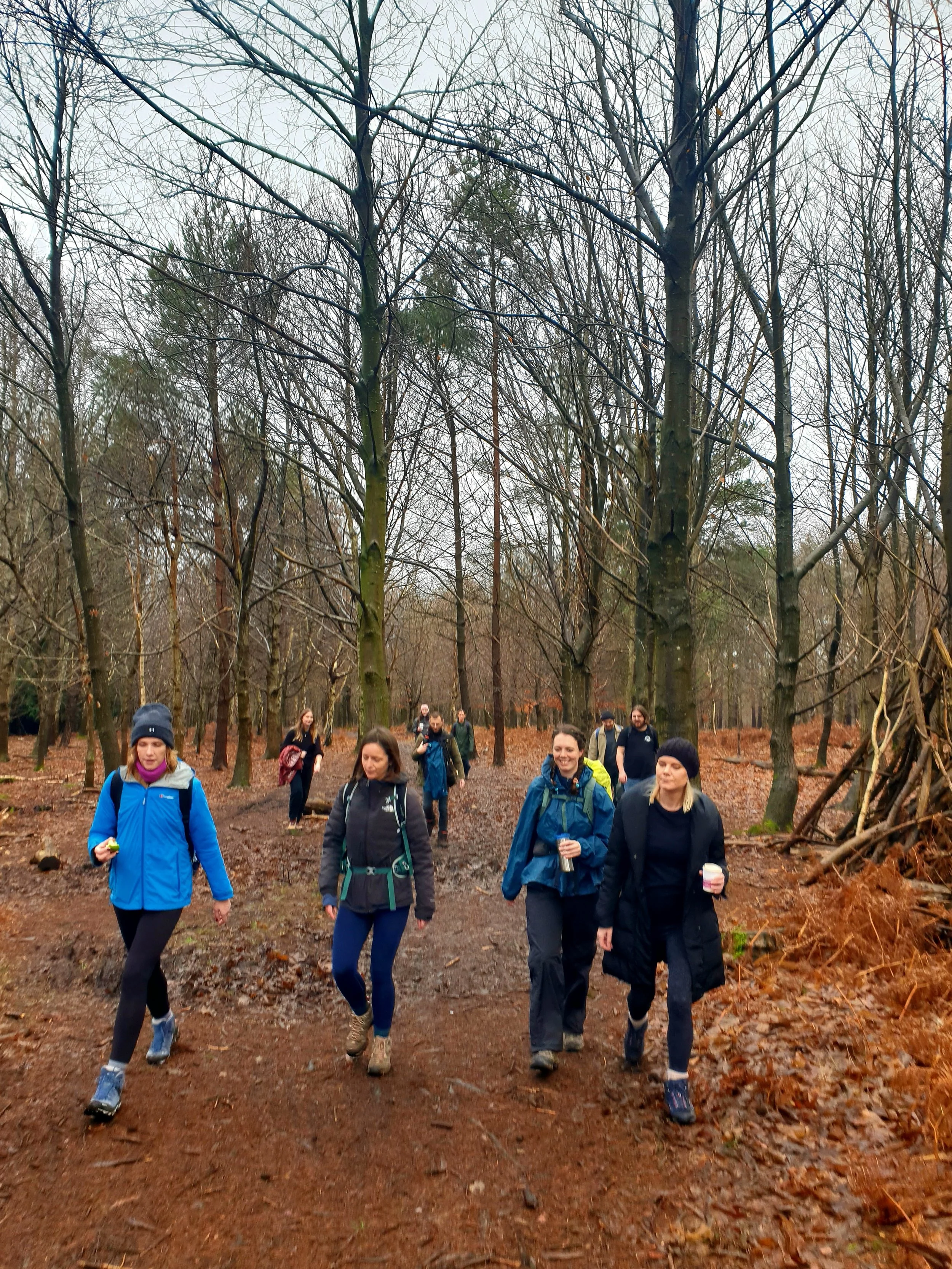 A group of women walking on a dirt trail through a forest with bare trees and fallen leaves.
