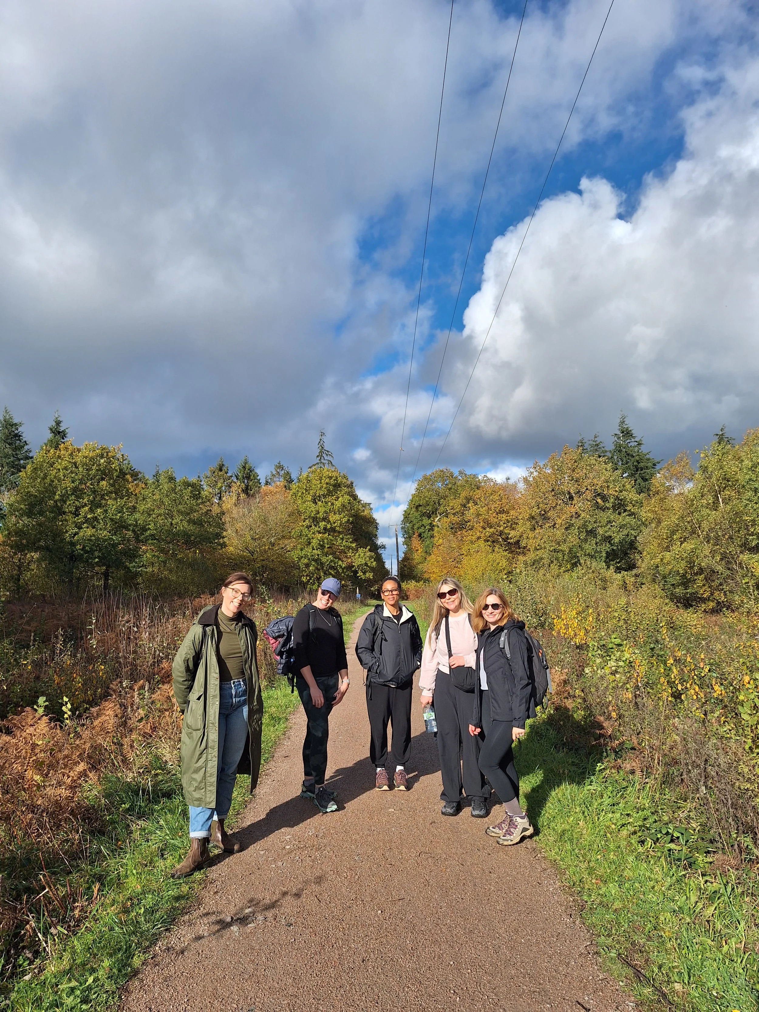 Five women stand on a dirt trail in a park with colorful autumn trees behind them. They are dressed in casual outdoor clothing, some with backpacks, and are smiling at the camera. The sky is partly cloudy with blue patches visible.