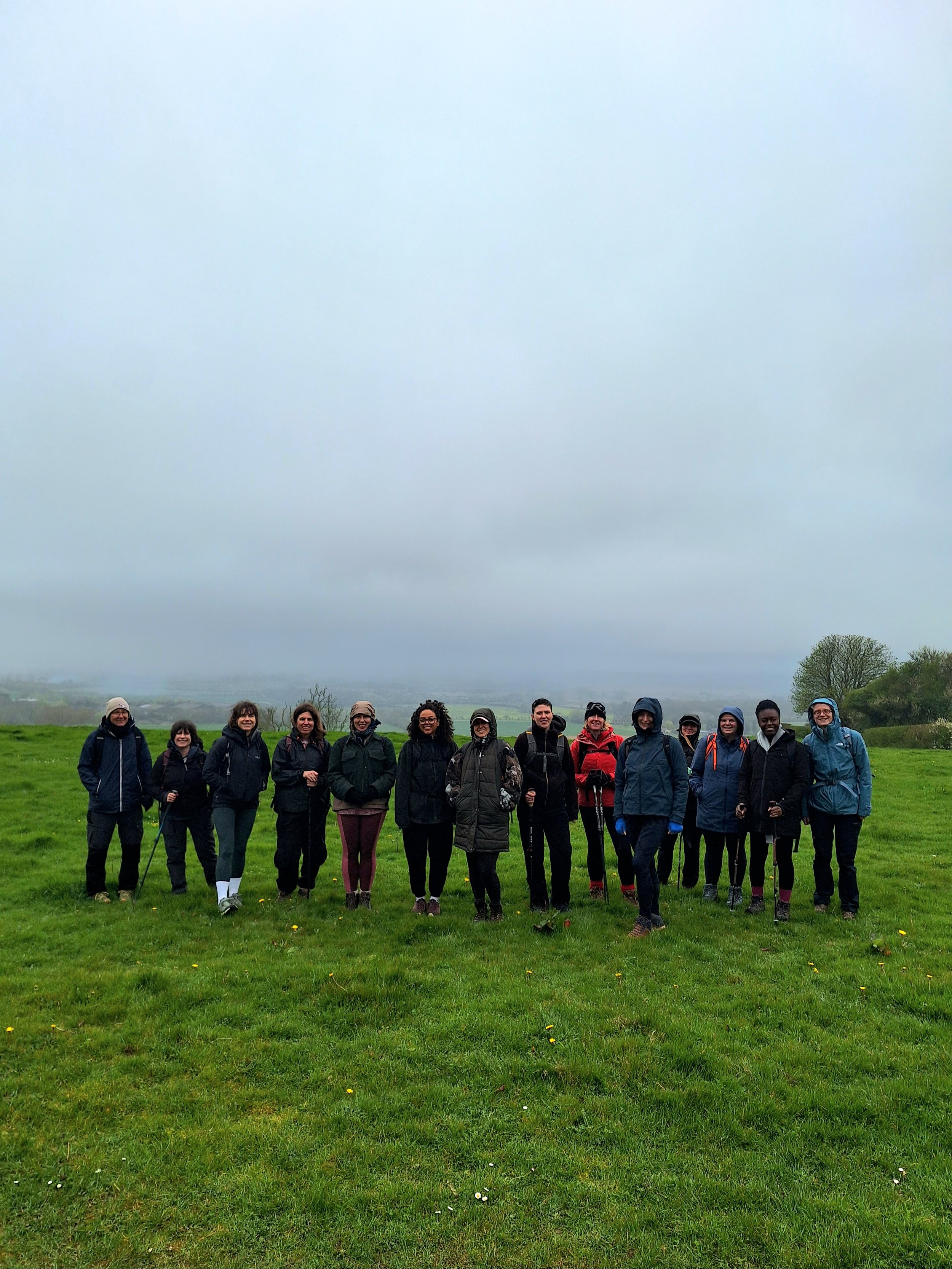 A group of women stands together outdoors on a grassy field with hills and trees in the background, wearing jackets and carrying backpacks on a cloudy day.