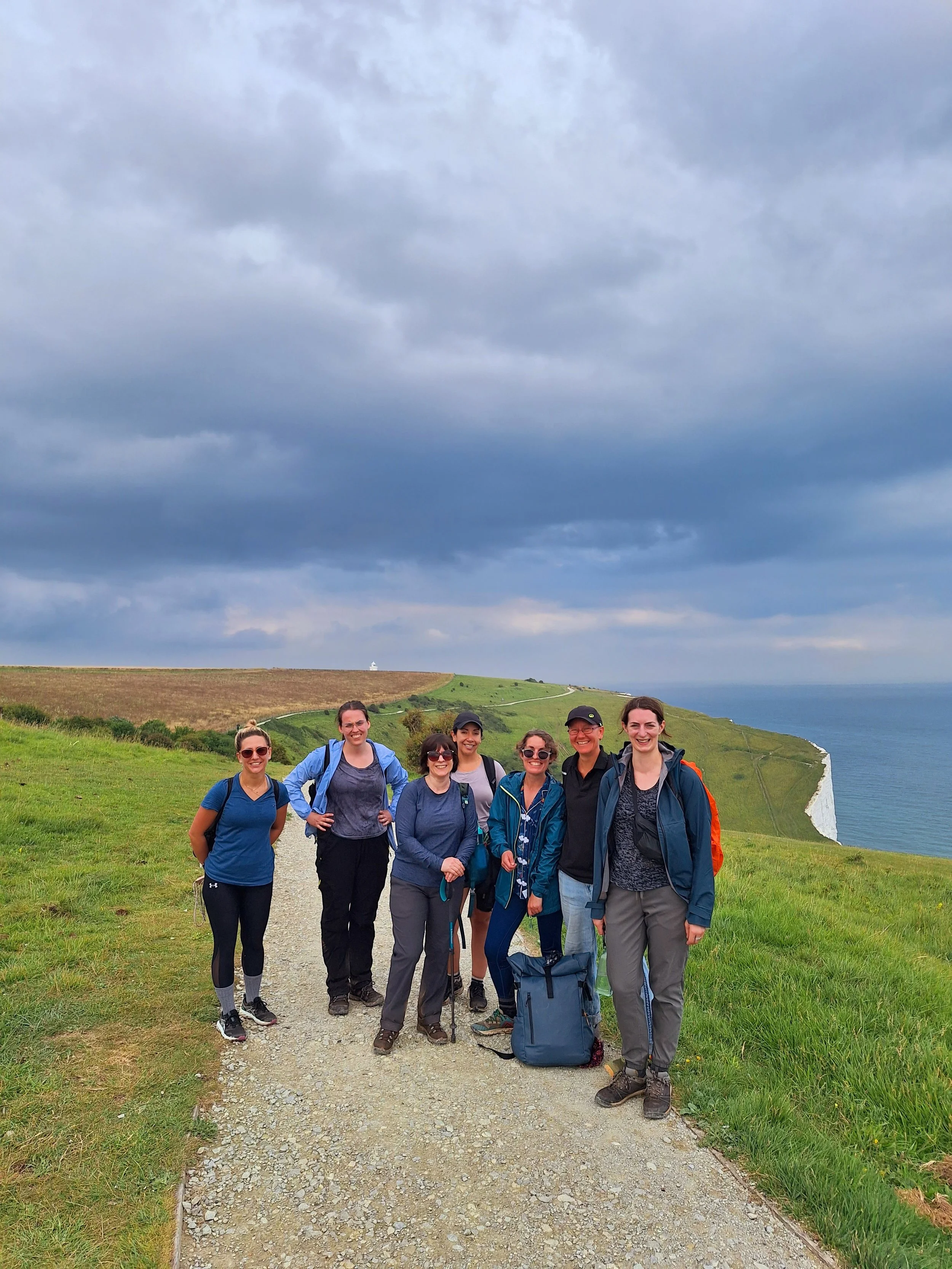 Group of seven women hikers standing on a dirt trail on a grassy hillside near the coast, with green fields, cliffs, and ocean under a cloudy sky in the background.