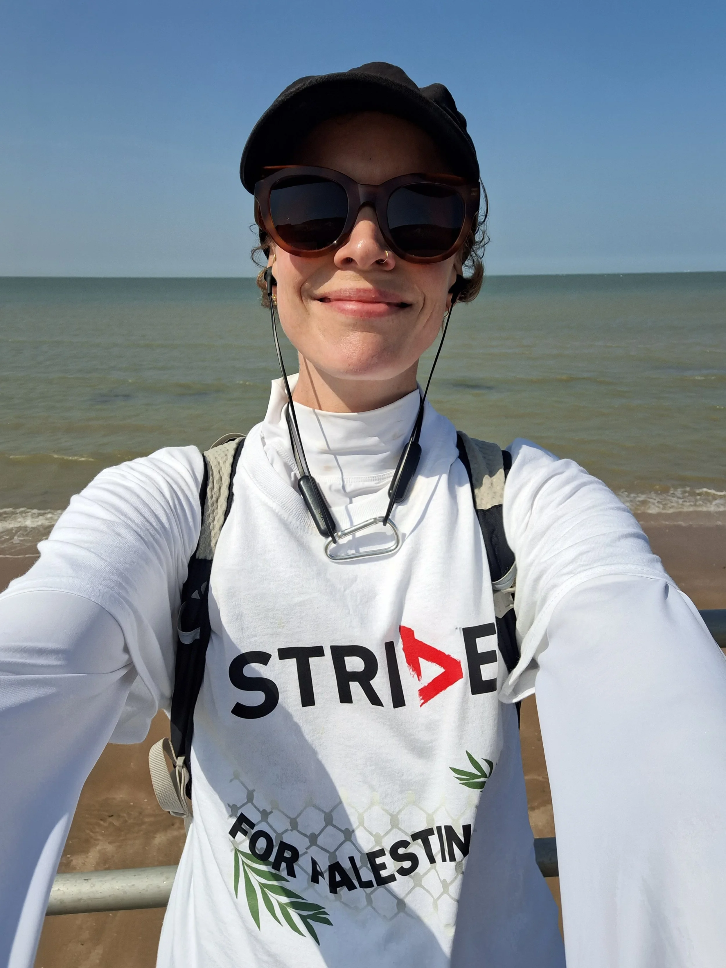 A woman taking a selfie in a cap wearing sportswear, smiling while standing in front of the sea