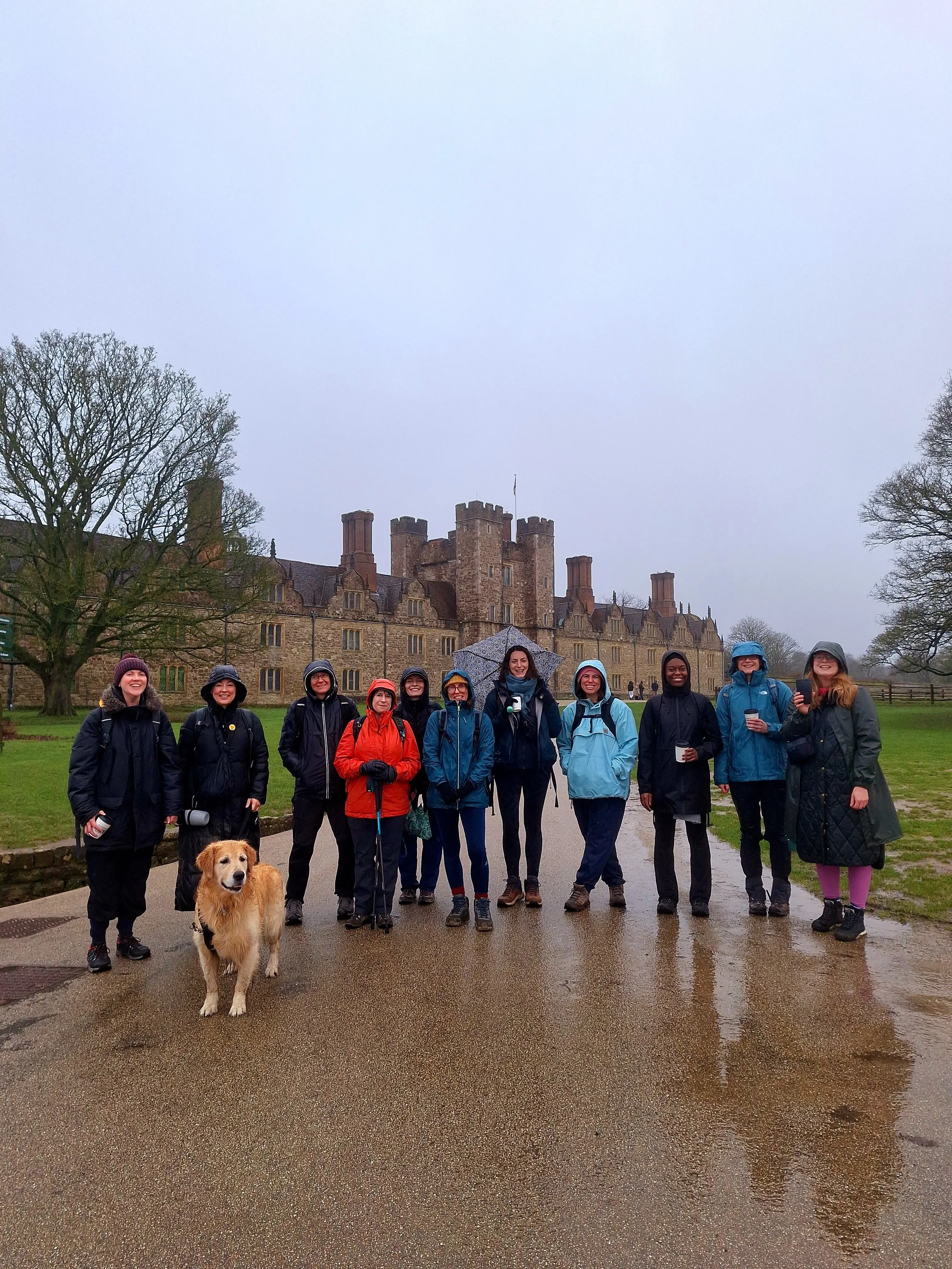 Group of women standing on a wet pathway outside a large historic stone building with turrets, holding umbrellas, with a dog in the foreground.