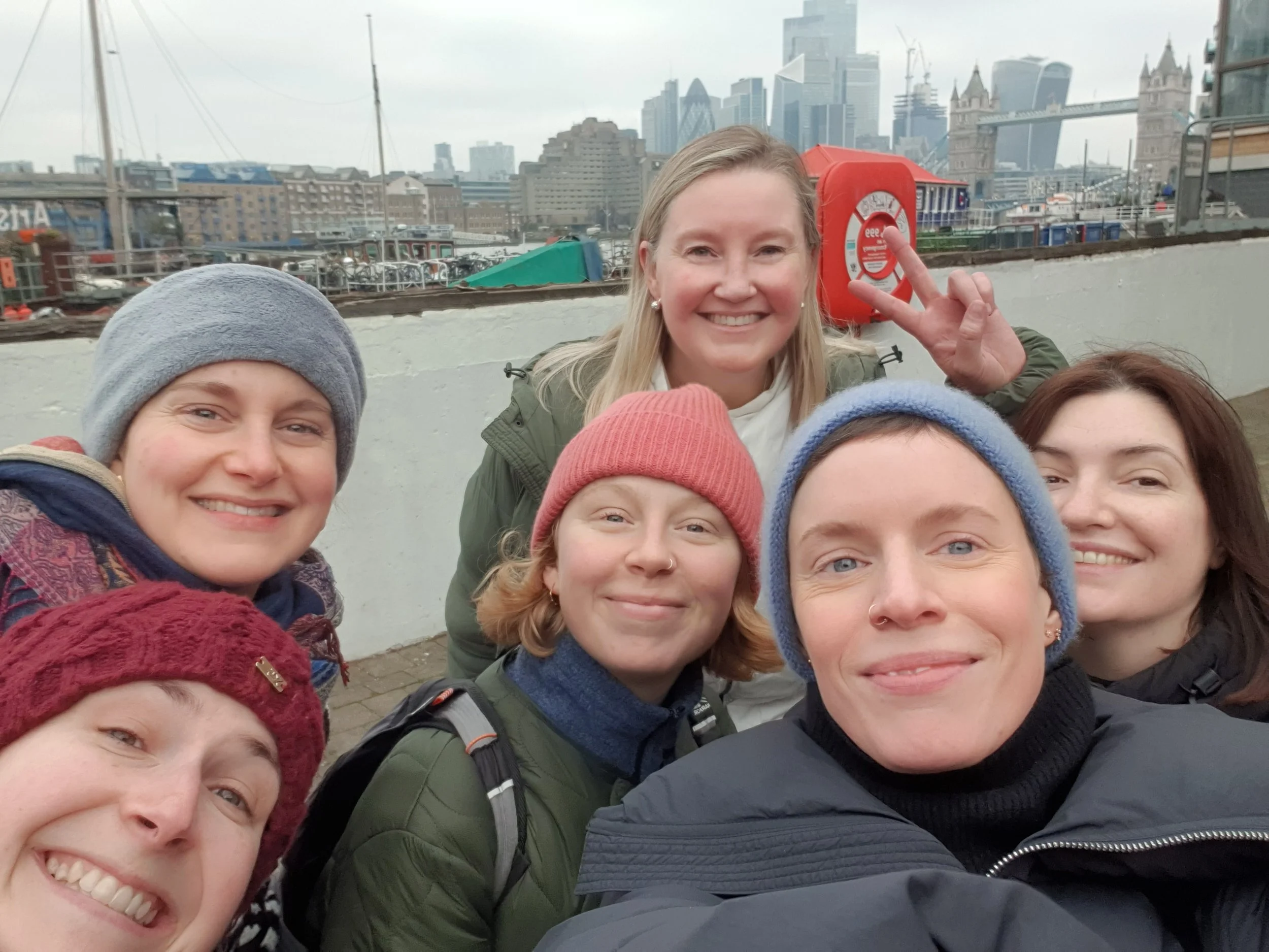 A group of smiling women posing closely together outdoors with the London skyline, including Tower Bridge, in the background.