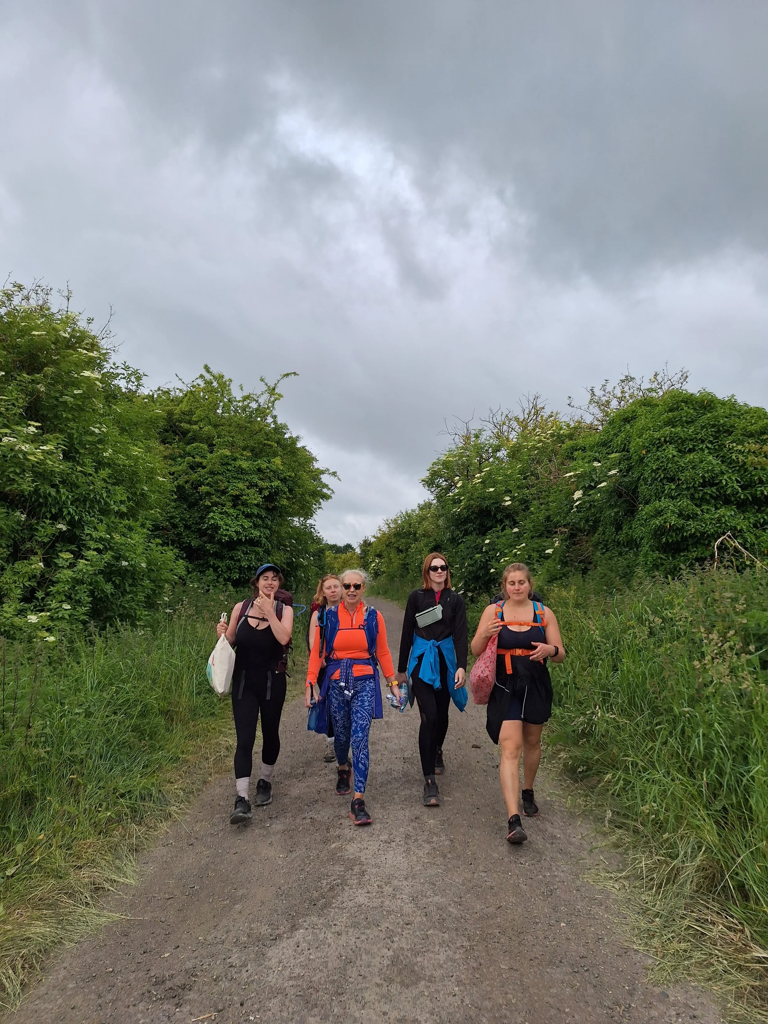 A group of five women walking along a dirt path surrounded by lush green trees and shrubs, with dark cloudy skies overhead.