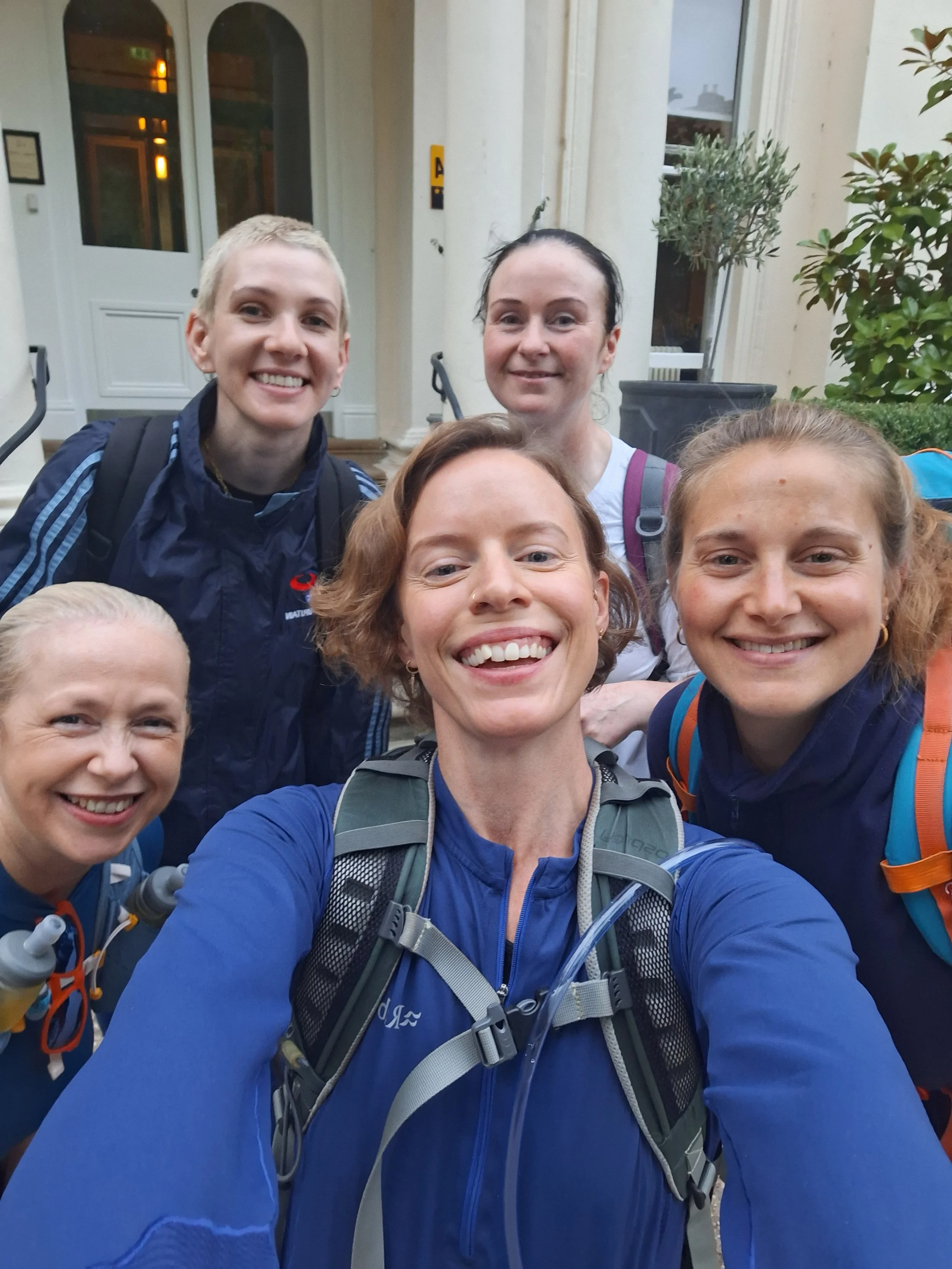 A group of five smiling women in sportswear, standing in front of a building doorway 