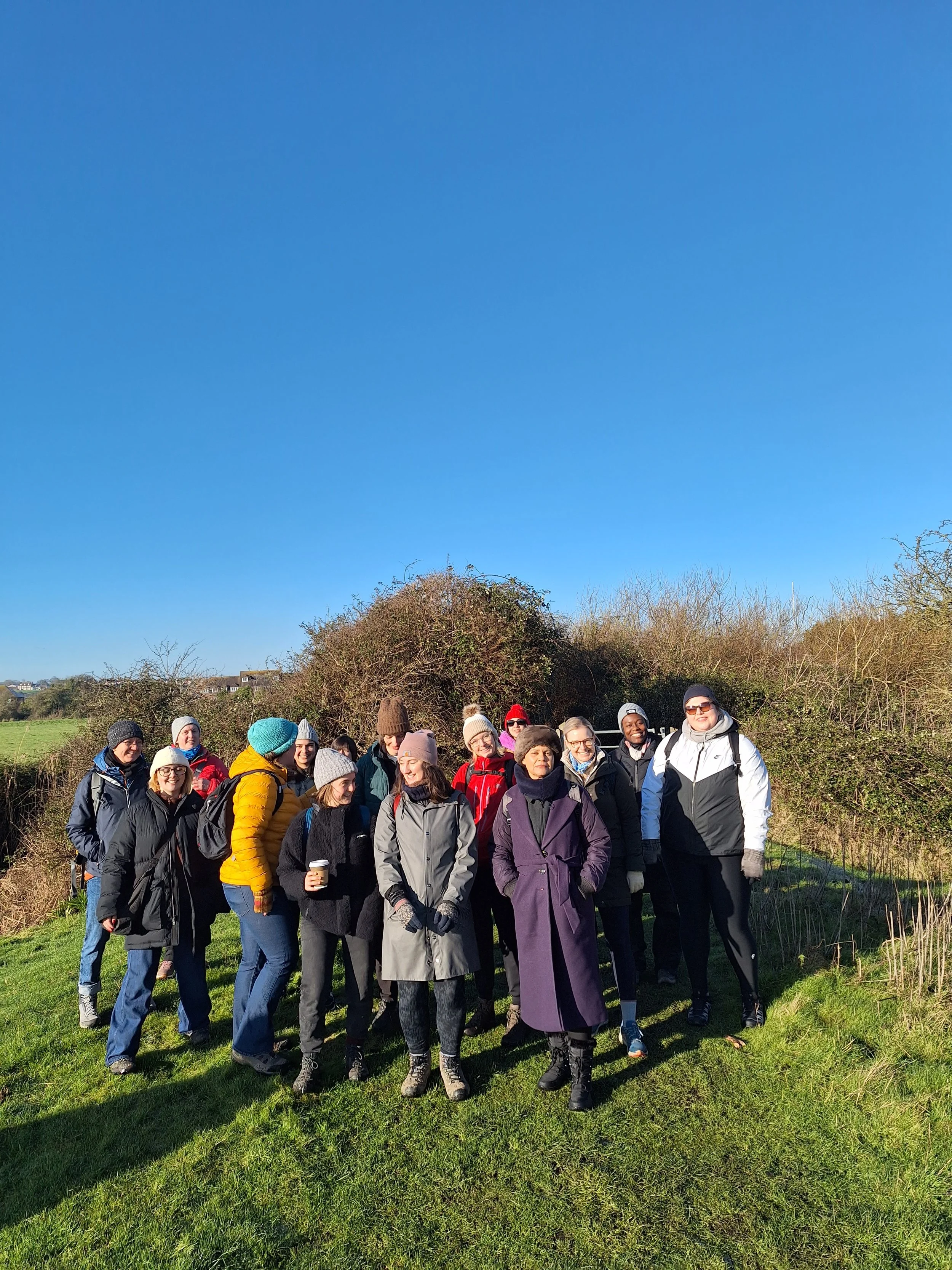 Group of diverse women standing outdoors on grass in a field with bushes and trees, dressed in warm clothing, enjoying a sunny day.