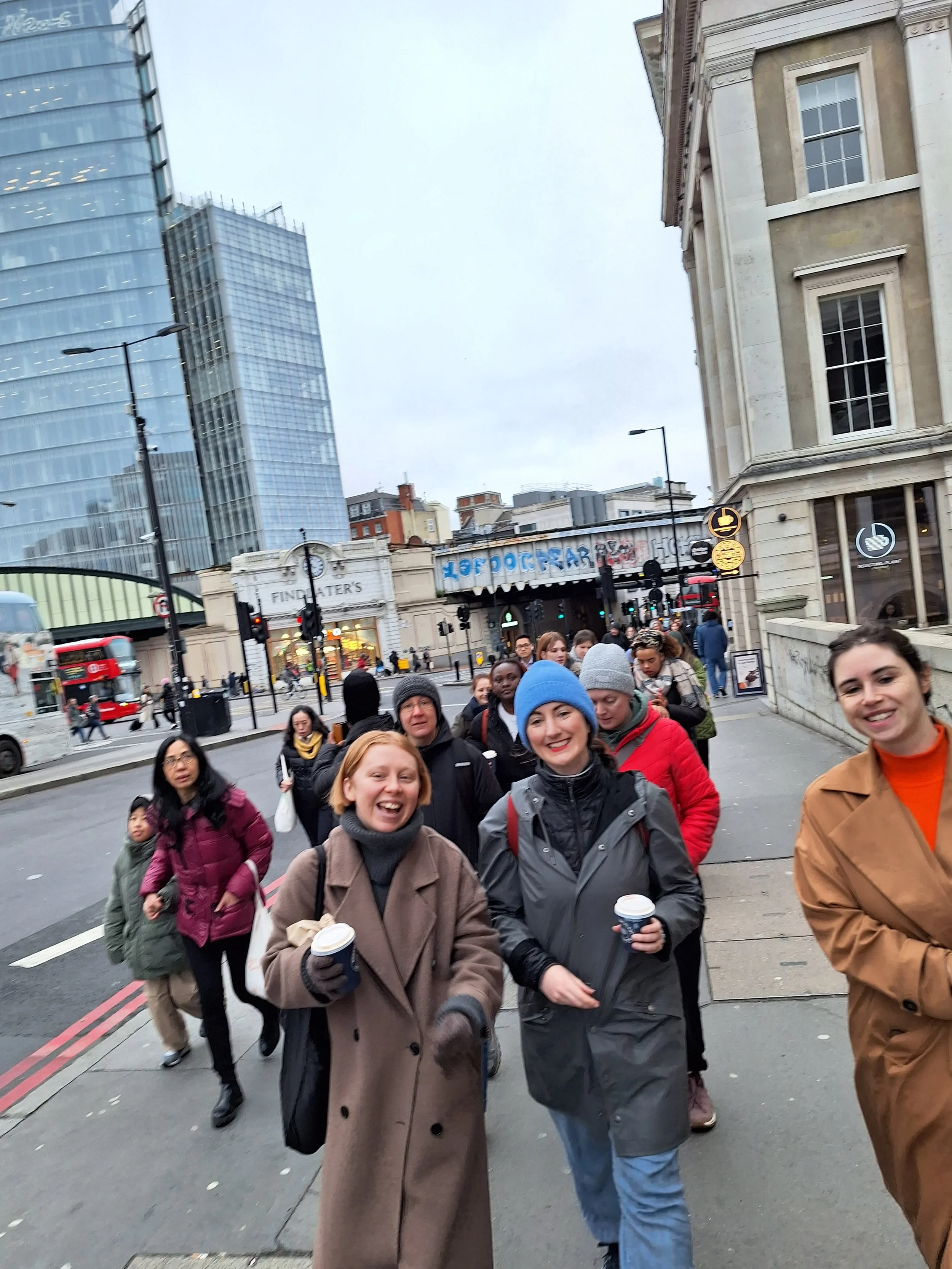 A group of smiling people walking on a city street holding coffee cups. Behind them are tall buildings, streetlights, and traffic signals.