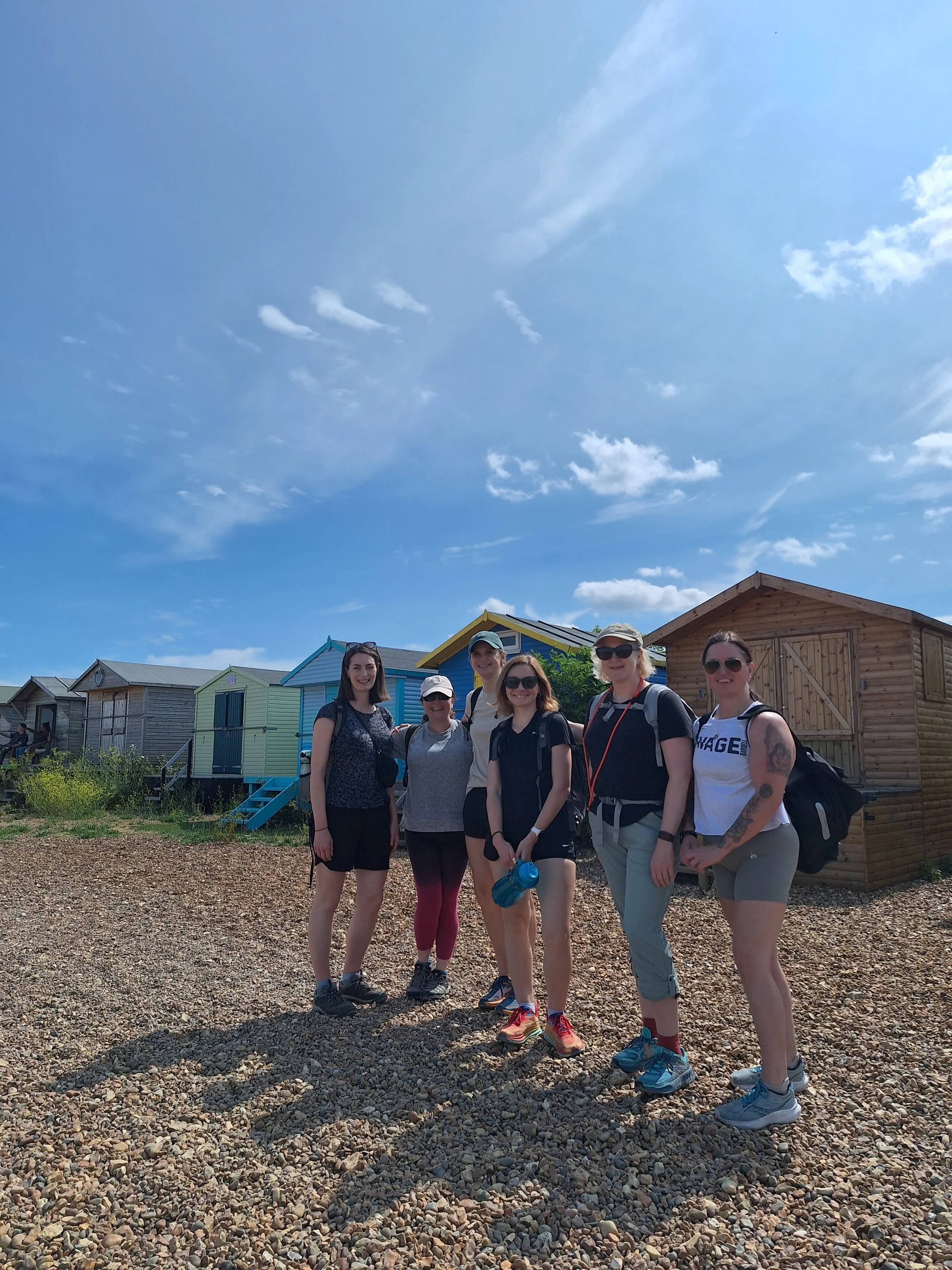 Group of six women standing outdoors on a sunny day in front of colorful beach huts, smiling at the camera.