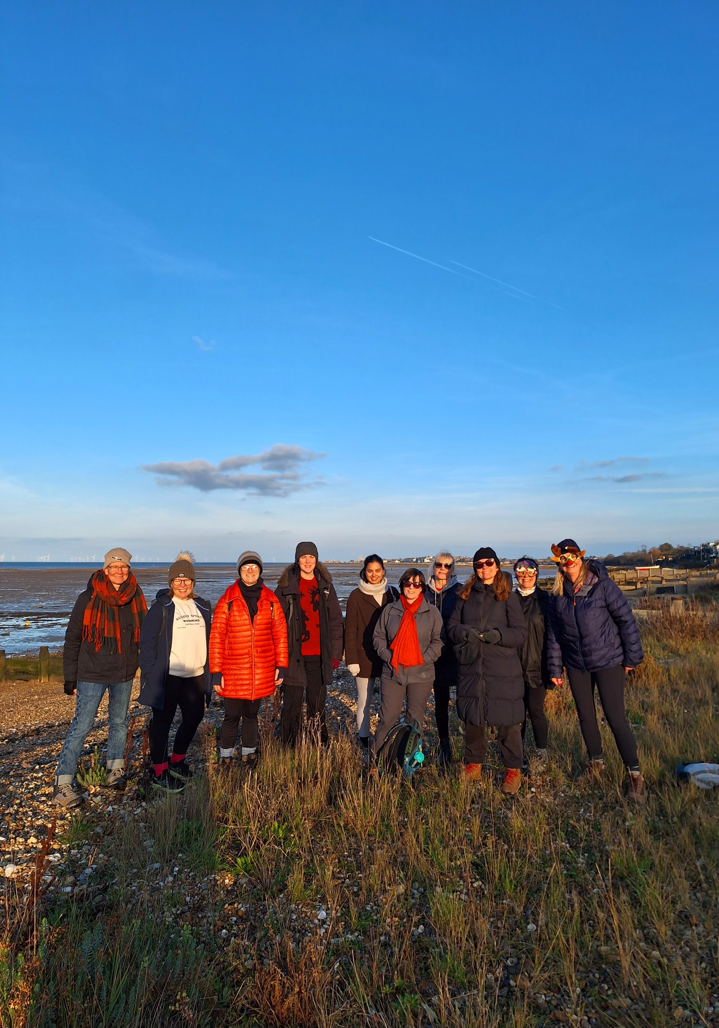 A group of ten women standing outdoors on a beach with a partly cloudy sky and contrails overhead. They are dressed in casual, layered clothing suitable for cool weather, and some are smiling at the camera.