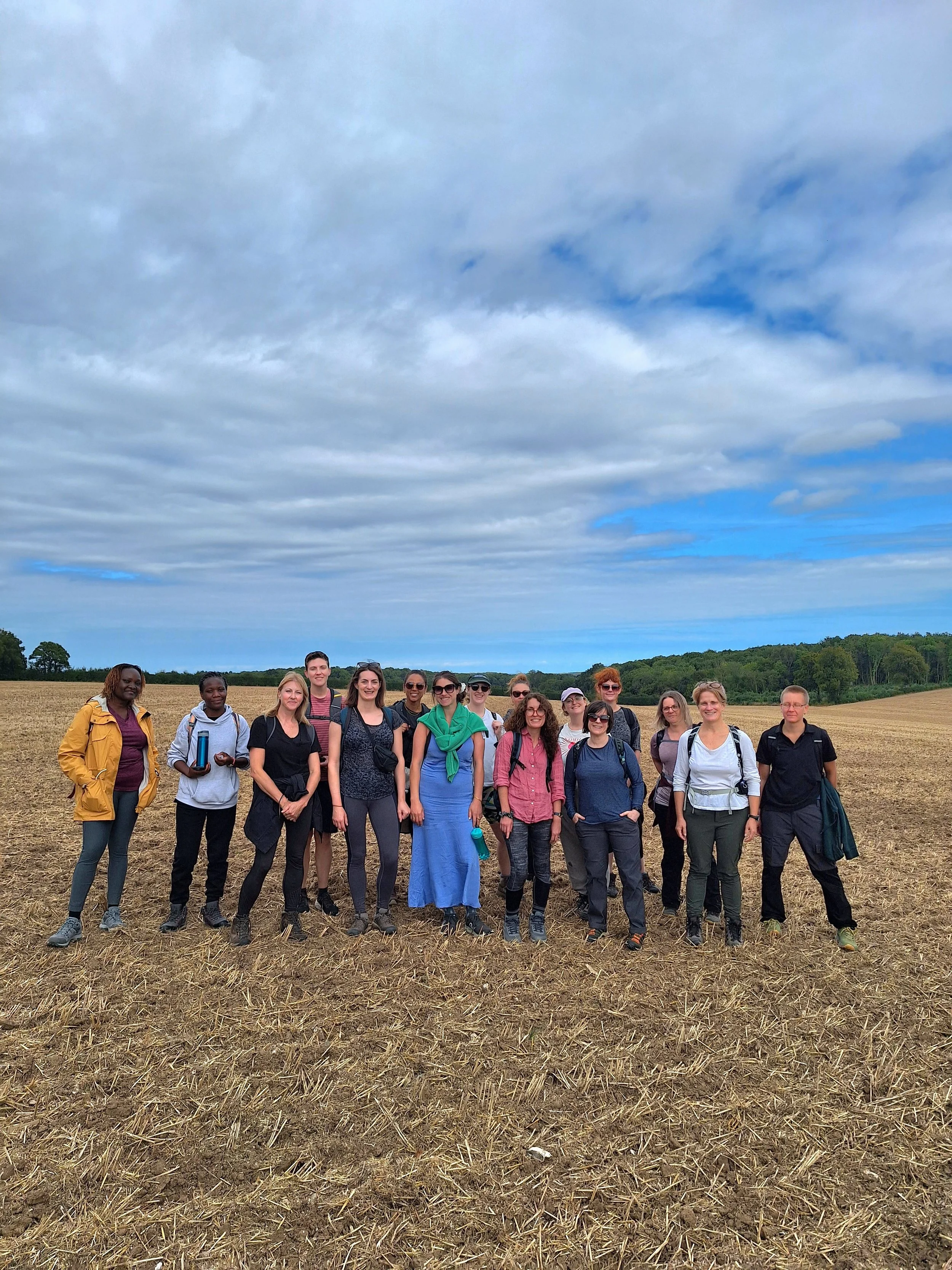 Group of women standing outdoors in a field with a cloudy sky above, dressed casually with backpacks and water bottles.