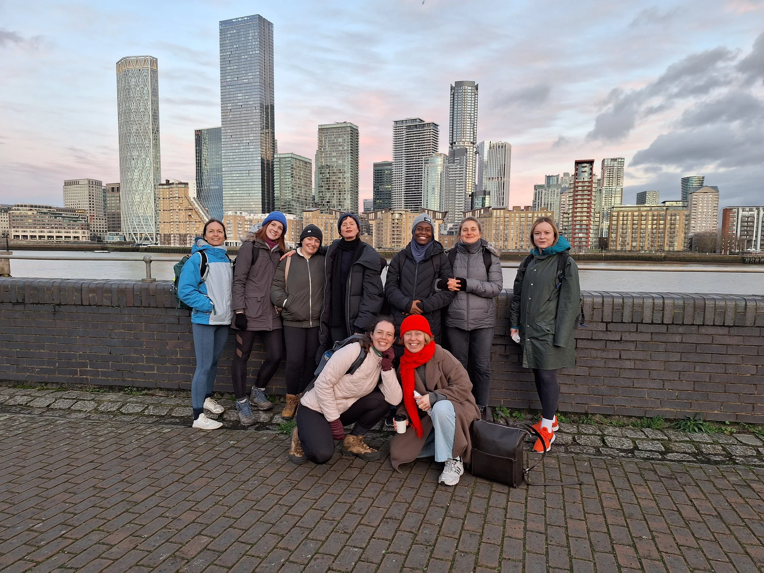 Group of eight women posing outdoors near a river with city skyscrapers in the background during cloudy weather.