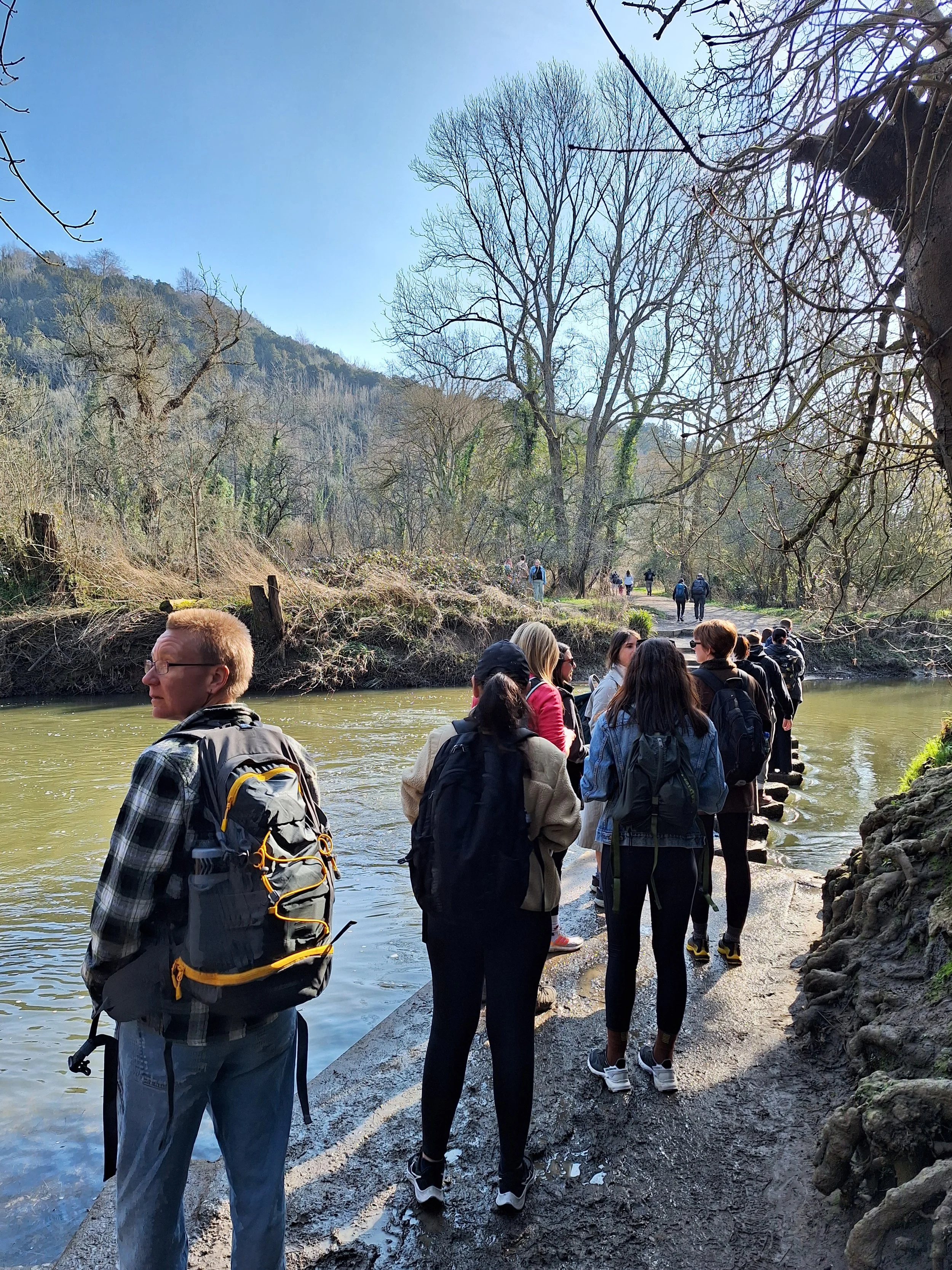 Group of women standing by a riverbank on a sunny day, surrounded by leafless trees and a hill in the background.