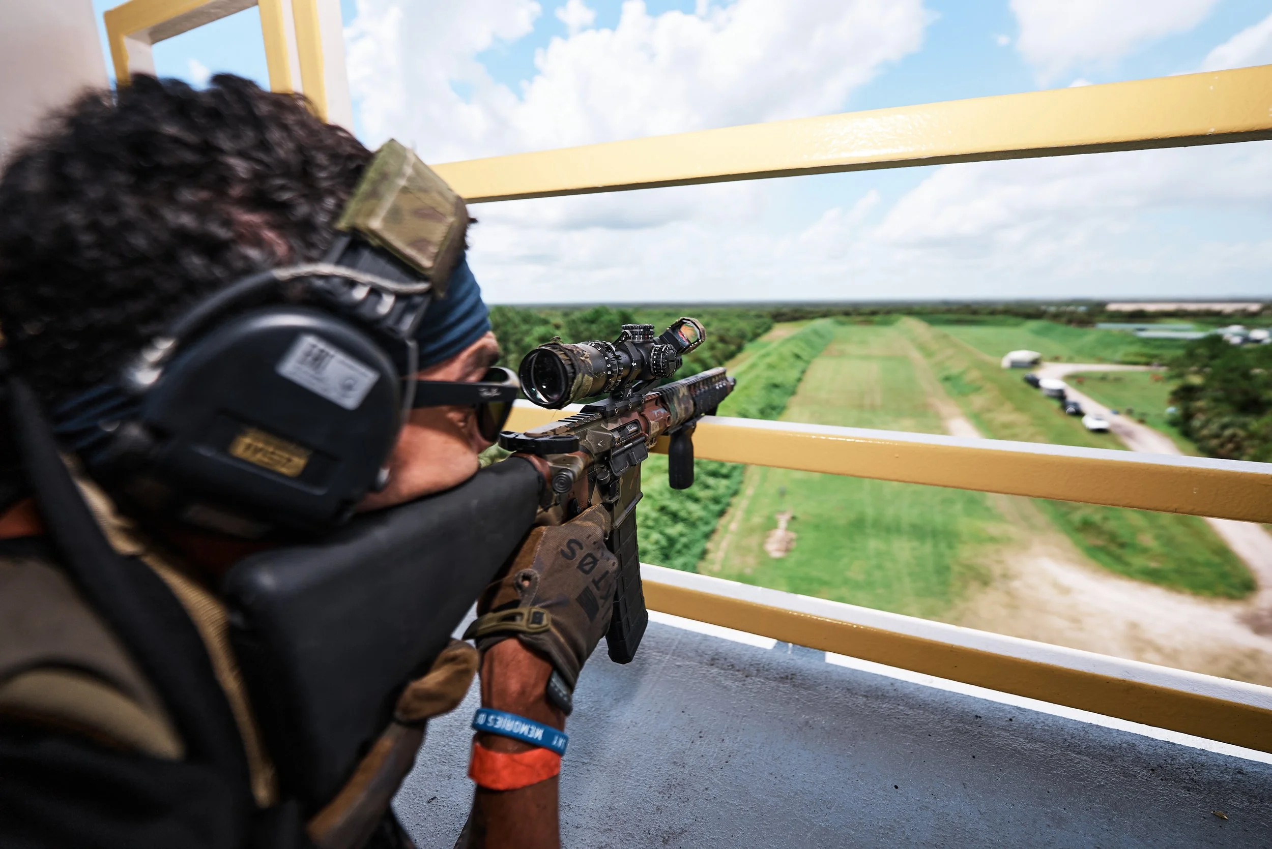 A person wearing headphones, sunglasses, and camouflage clothing, aiming a rifle with a scope from a tower or elevated position over a green field with trees and a gravel path, under a partly cloudy sky.