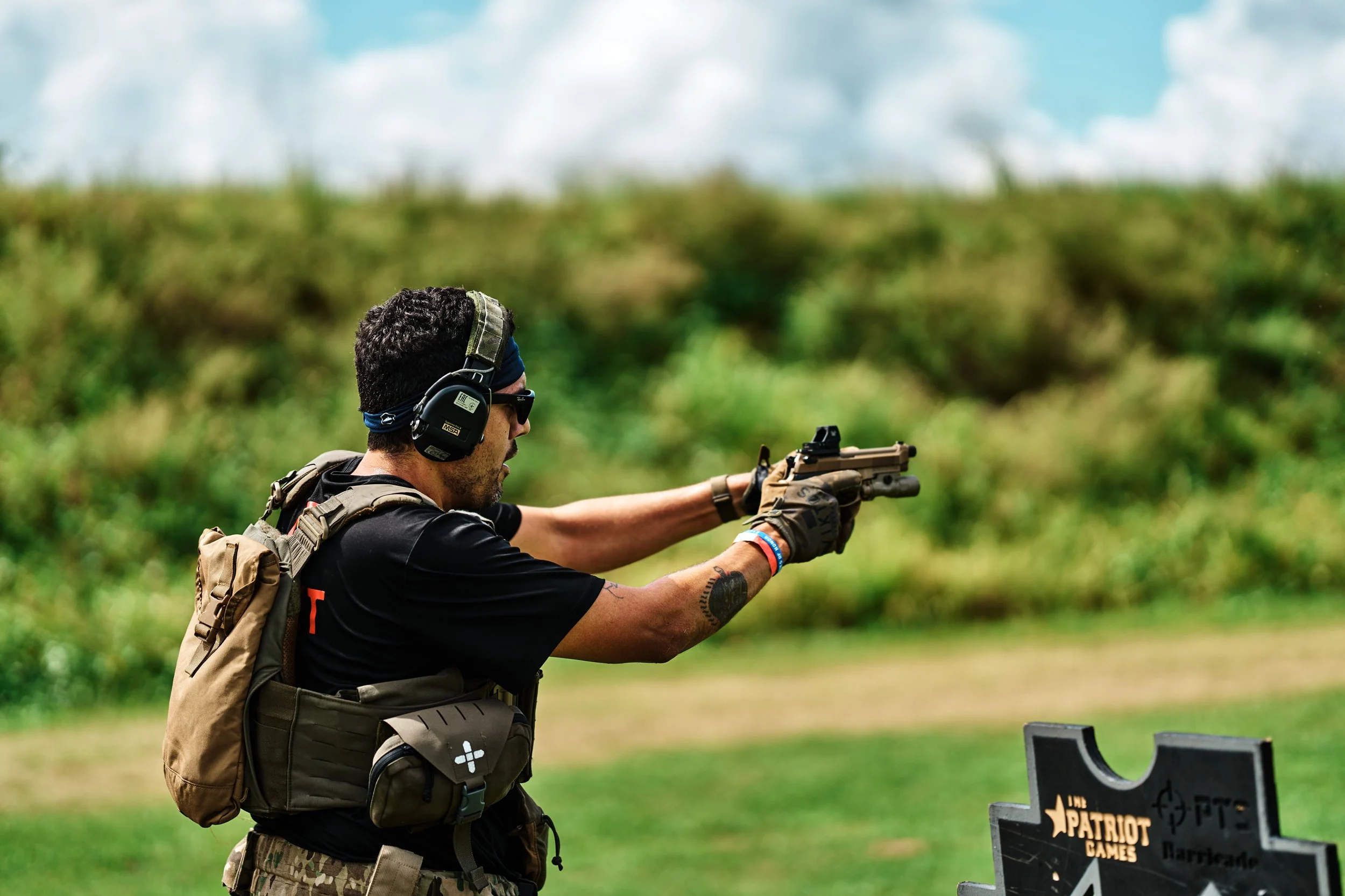 A man aiming a handgun at a target outdoors, wearing protective headphones and sunglasses, with a backpack and tactical gear.