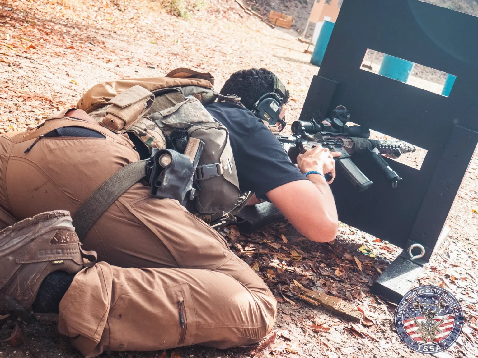 A person lying prone behind a shooting barrier, aiming a rifle at a target, with tactical gear and a helmet, on a dirt shooting range with leaves on the ground and the logo of the Tactical Shooting Sports Association (TSSA) visible.
