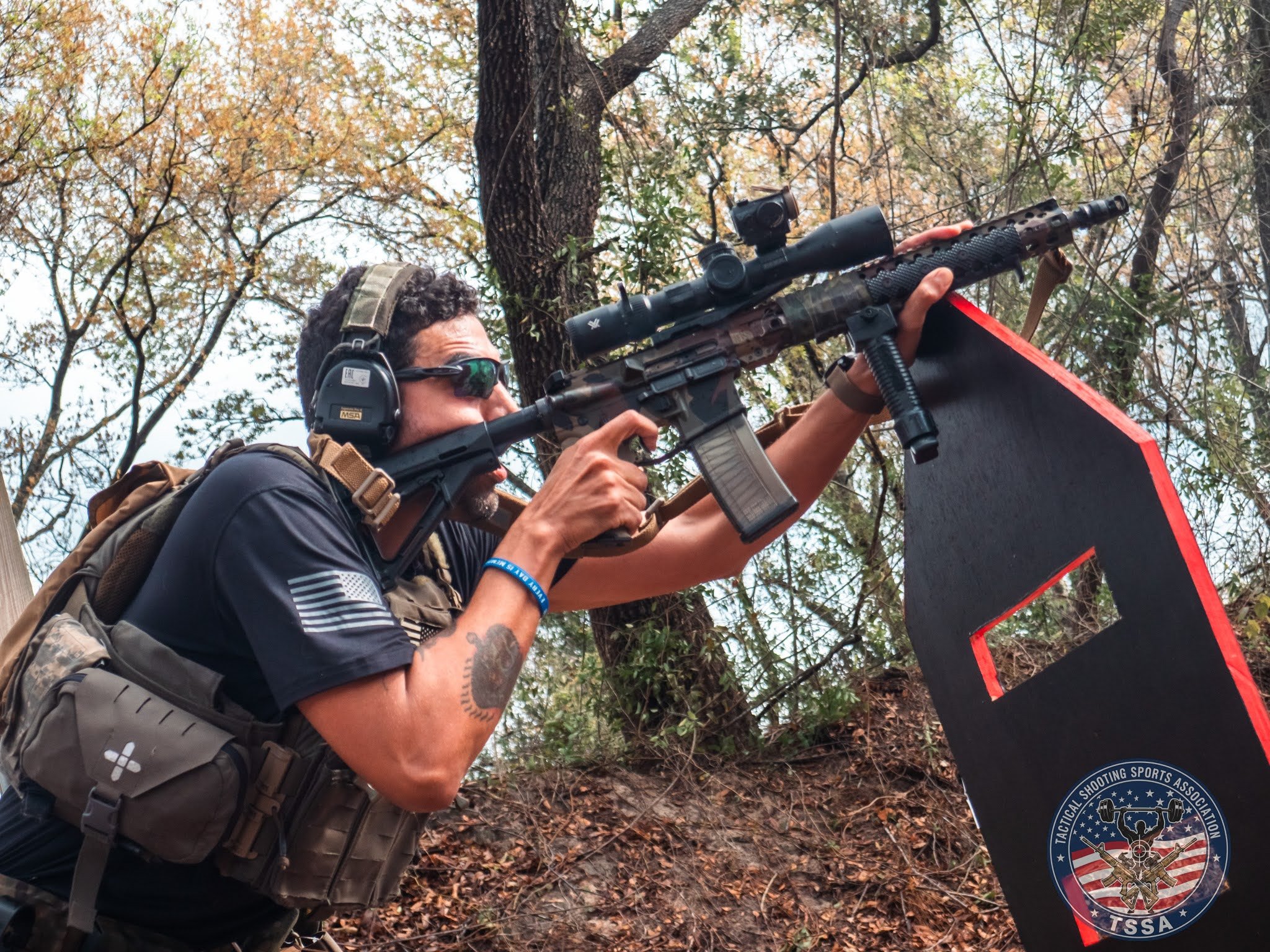 A man wearing tactical gear, sunglasses, and ear protection aiming a rifle on a shooting range in a wooded area.