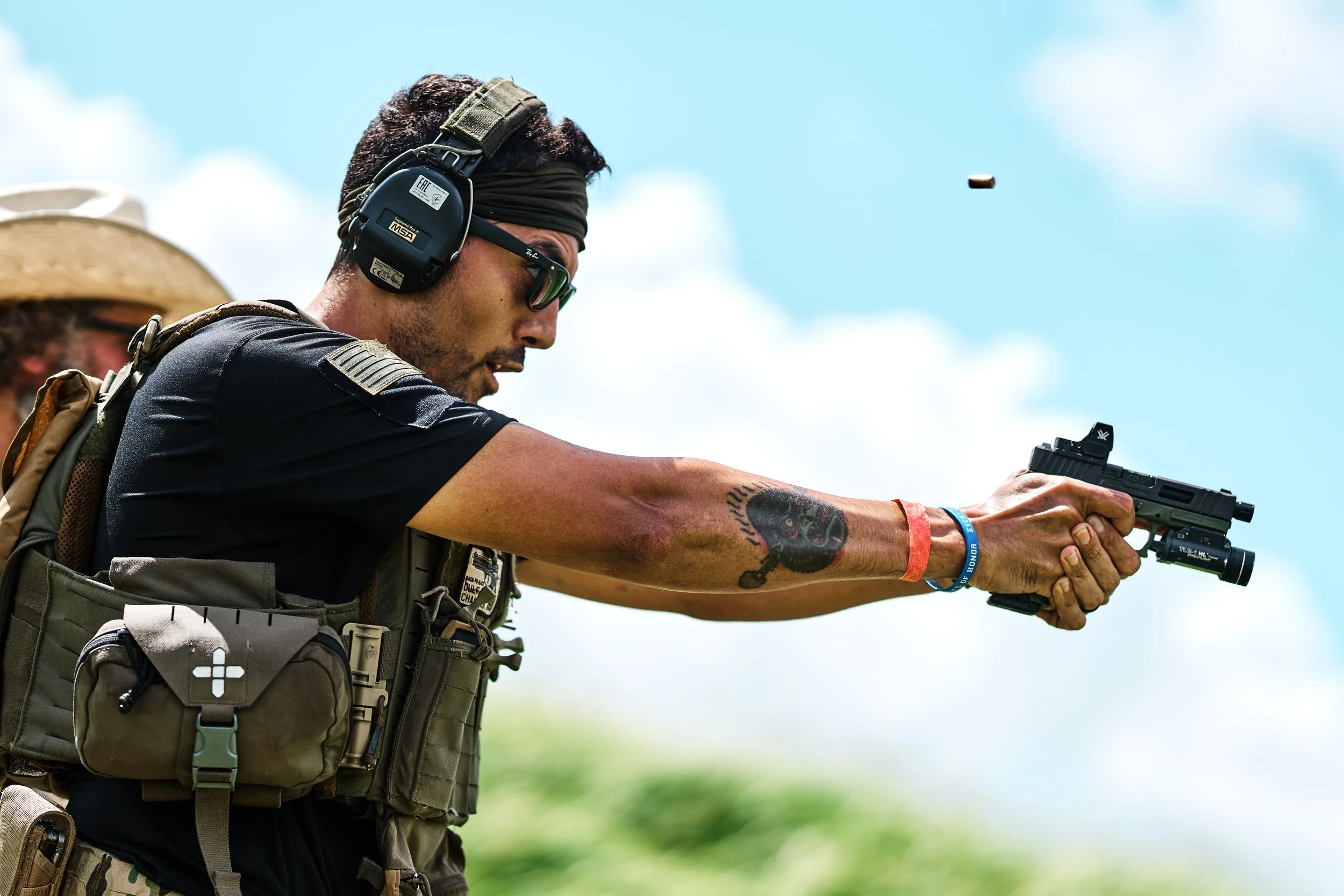 A man wearing sunglasses, a headband, and ear protection, aiming a handgun at a target outdoors, with another person partially visible in the background.