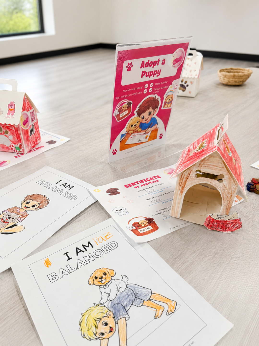 Flat lay of Puppy Yoga camp materials including Adopt a Puppy sign, handmade doghouses, Certificate of Adoption, and I Am Balanced affirmation pages at The Mindfulness Movement retreat
