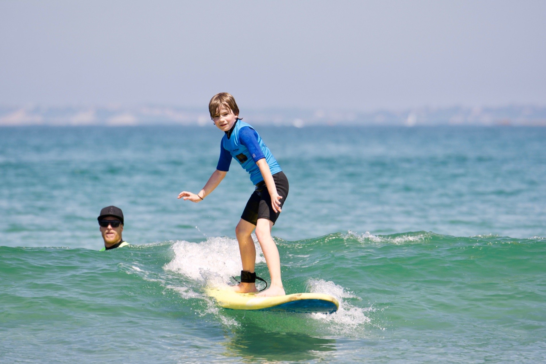Niño surfeando en una tabla amarilla en el mar, con instructor observando en el agua.