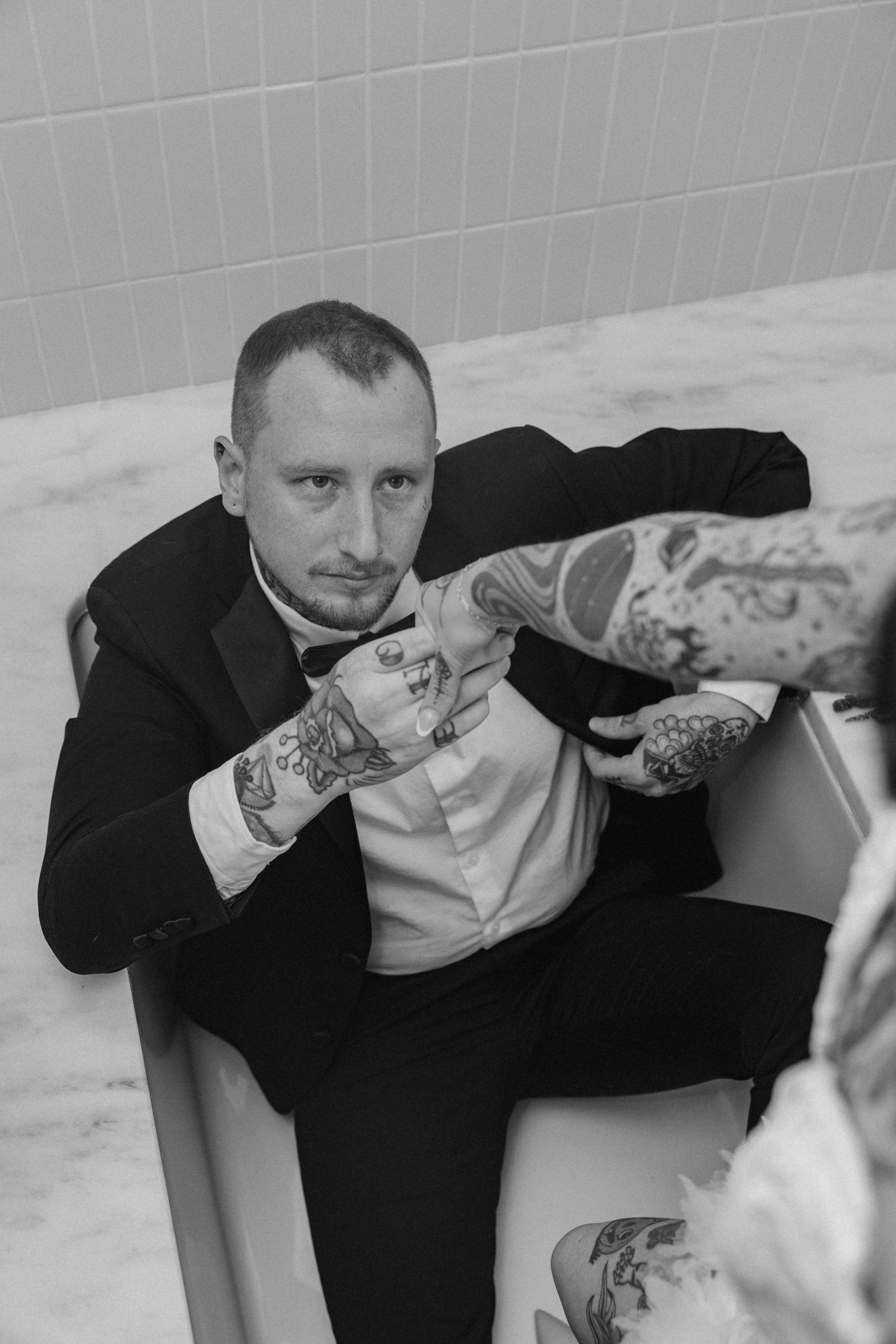 A man in a black tuxedo with a bowtie, seated in a bathtub, pointing a finger at someone outside the frame. He has tattoos on his arms and hands, with a serious expression, in a monochrome photo.