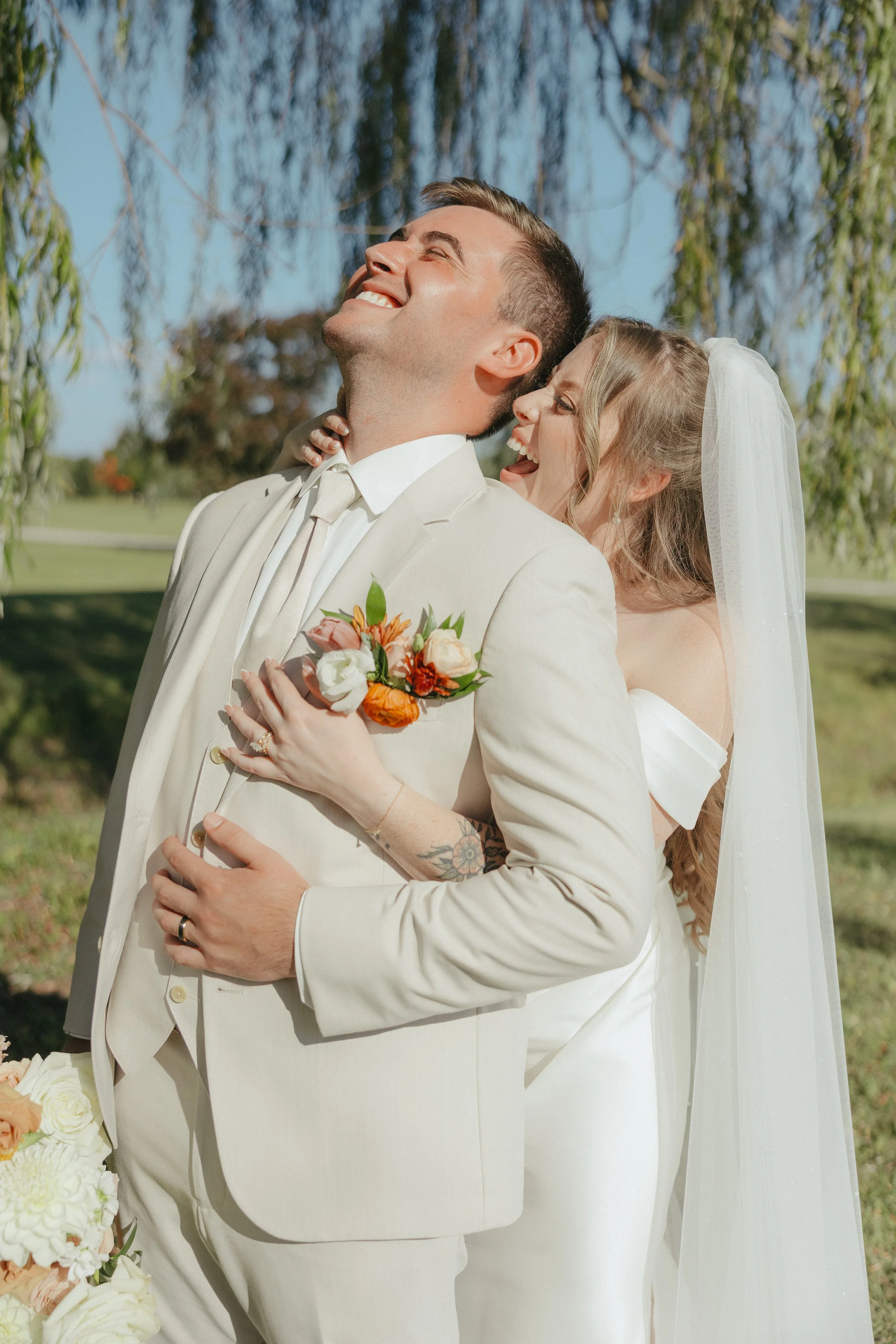A joyful couple on their wedding day outdoors, embracing and smiling happily, with trees and a clear blue sky in the background.