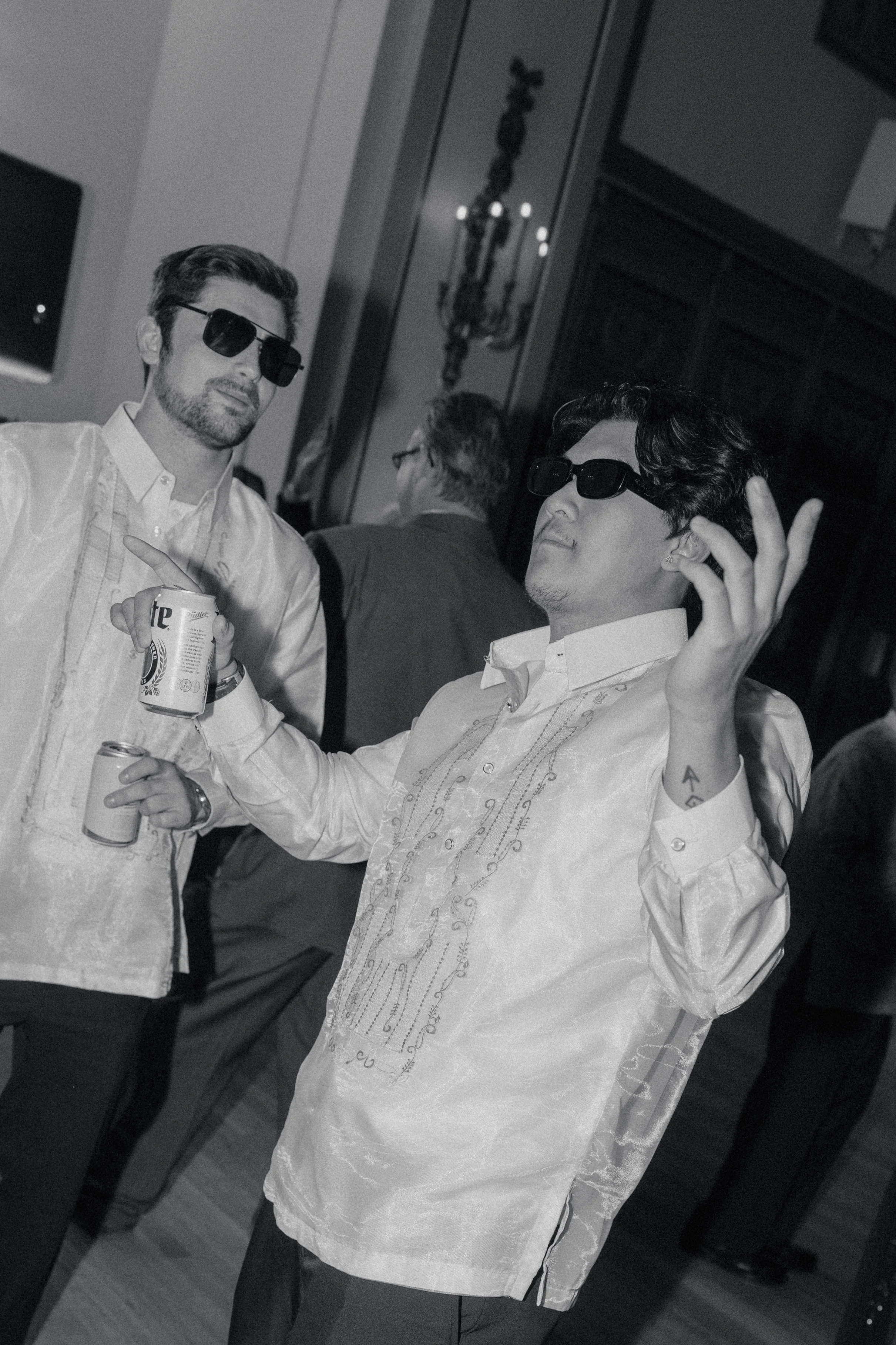 Two men wearing traditional Barong Tagalog shirts and sunglasses, holding drinks, are posing and smiling at a social gathering. One man is gesturing with his hand while the other looks on.