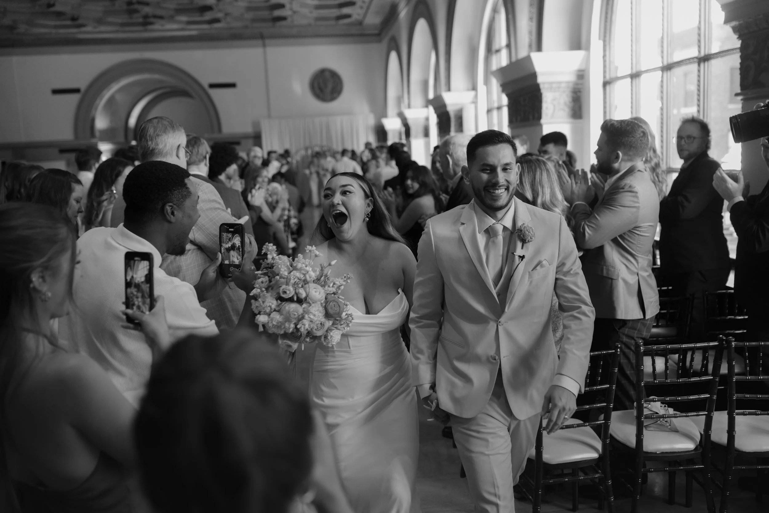 A black and white photo of a newlywed couple walking down the aisle, smiling and holding hands, with wedding guests taking photos and clapping.