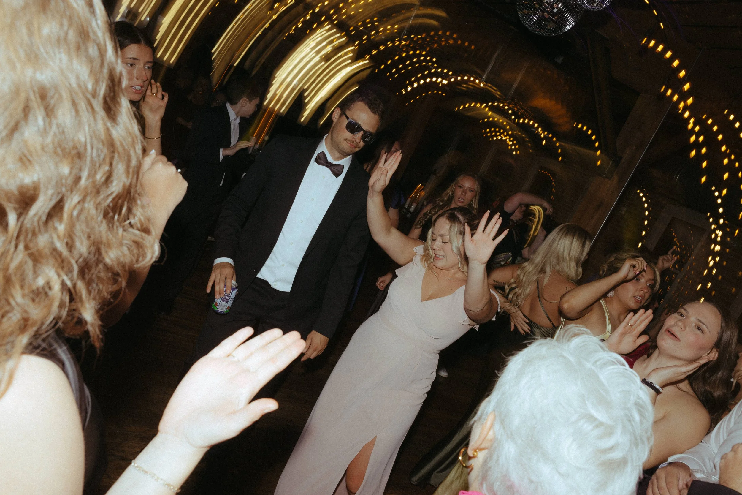 People dancing and enjoying themselves at a party or celebration in a dimly lit venue with string lights and a mirror wall.