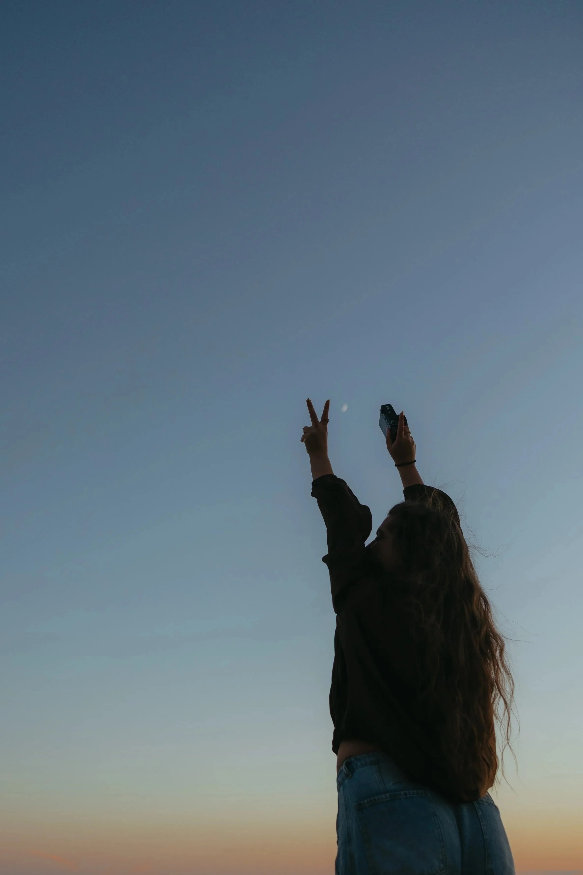 women holding camera and a peace sign up in front of moon and sunset