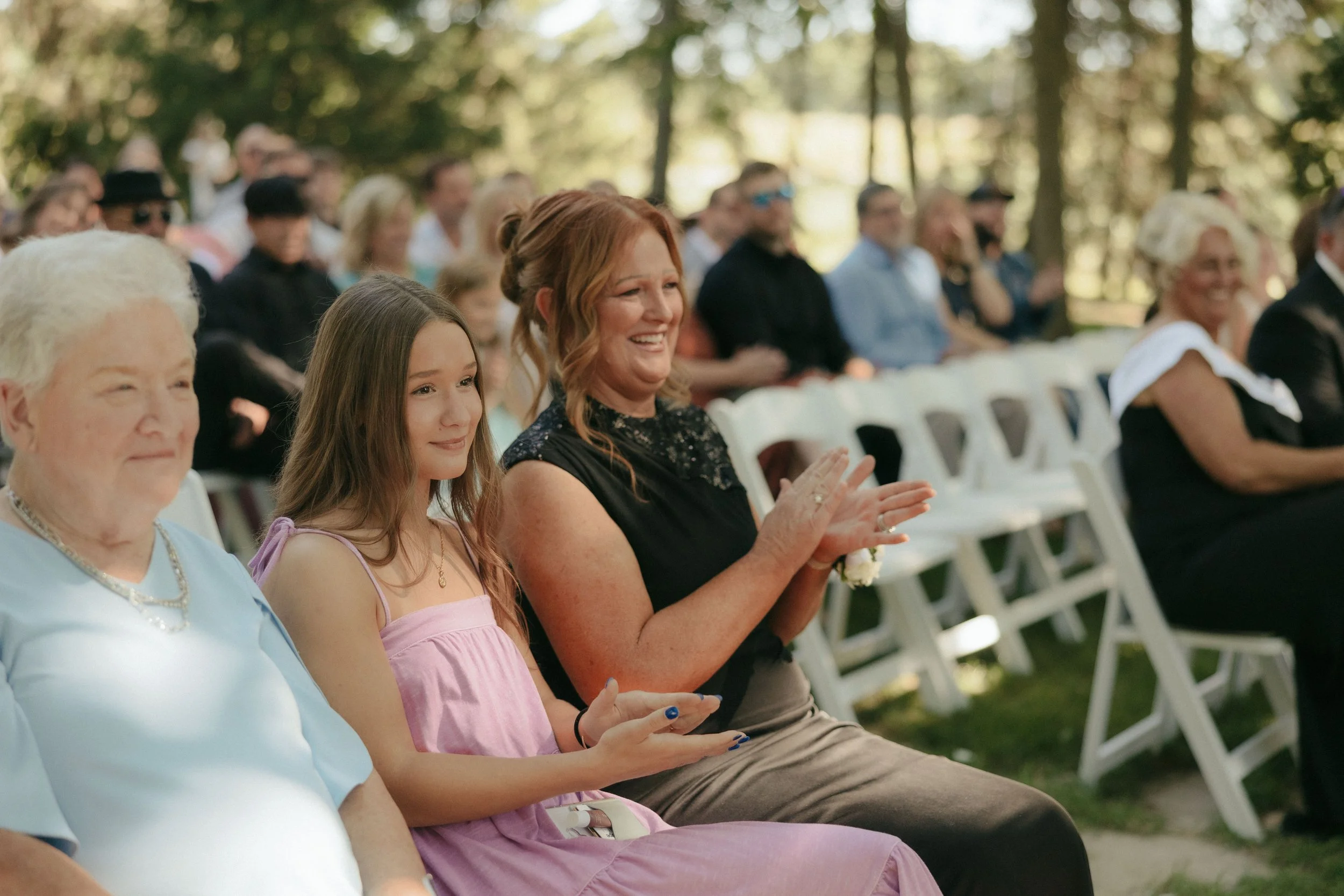 Group of people sitting outdoors at a wedding ceremony, some clapping and smiling, with trees in the background.