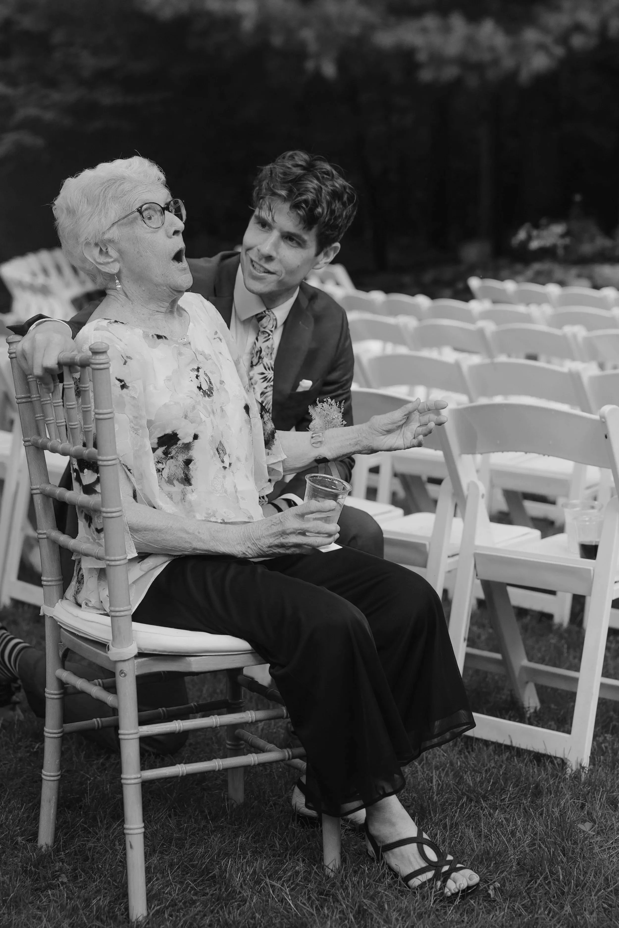 An elderly woman with glasses and a floral blouse sitting on a wooden chair with a young man in a blazer and patterned shirt, engaging in conversation at an outdoor event with empty chairs in the background.