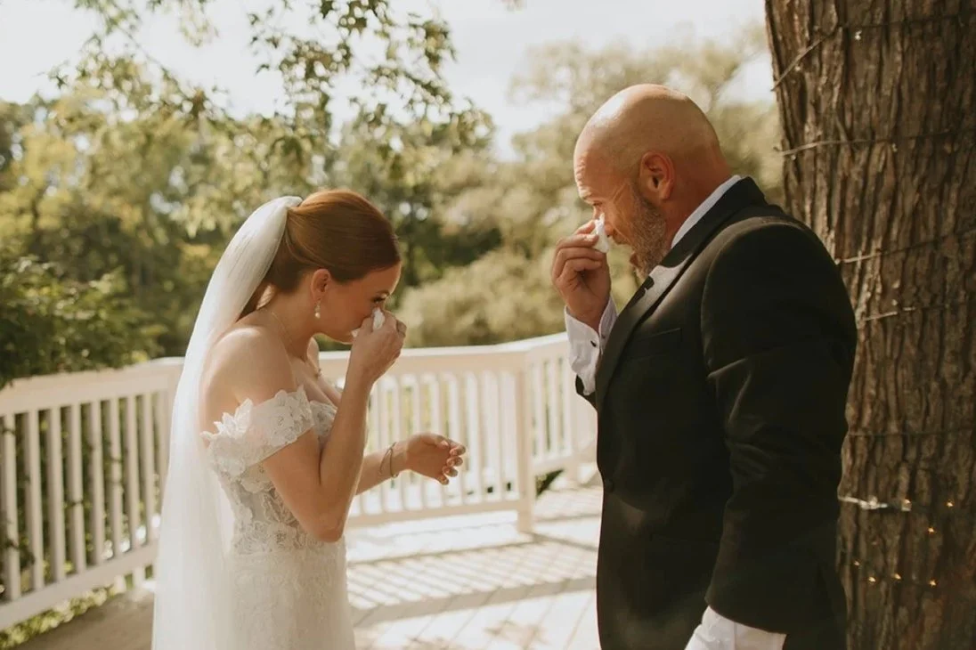 A bride and a groom crying tears of joy while holding tissues outdoors on a wooden porch with trees in the background.