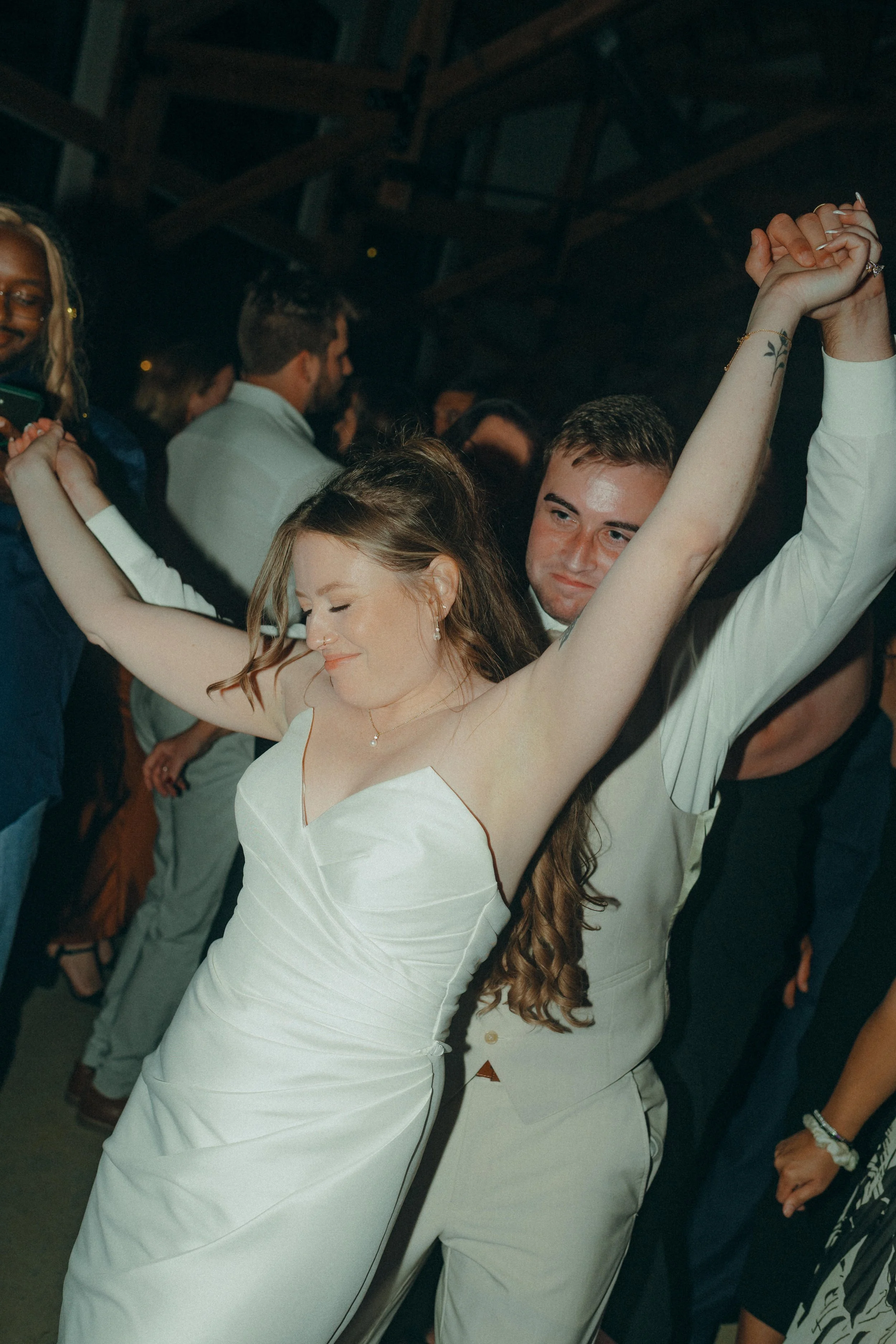 Couple dancing at a celebration, the woman in a white dress with arms raised, smiling with eyes closed, the man standing behind her with arms around her, in a lively indoor setting.