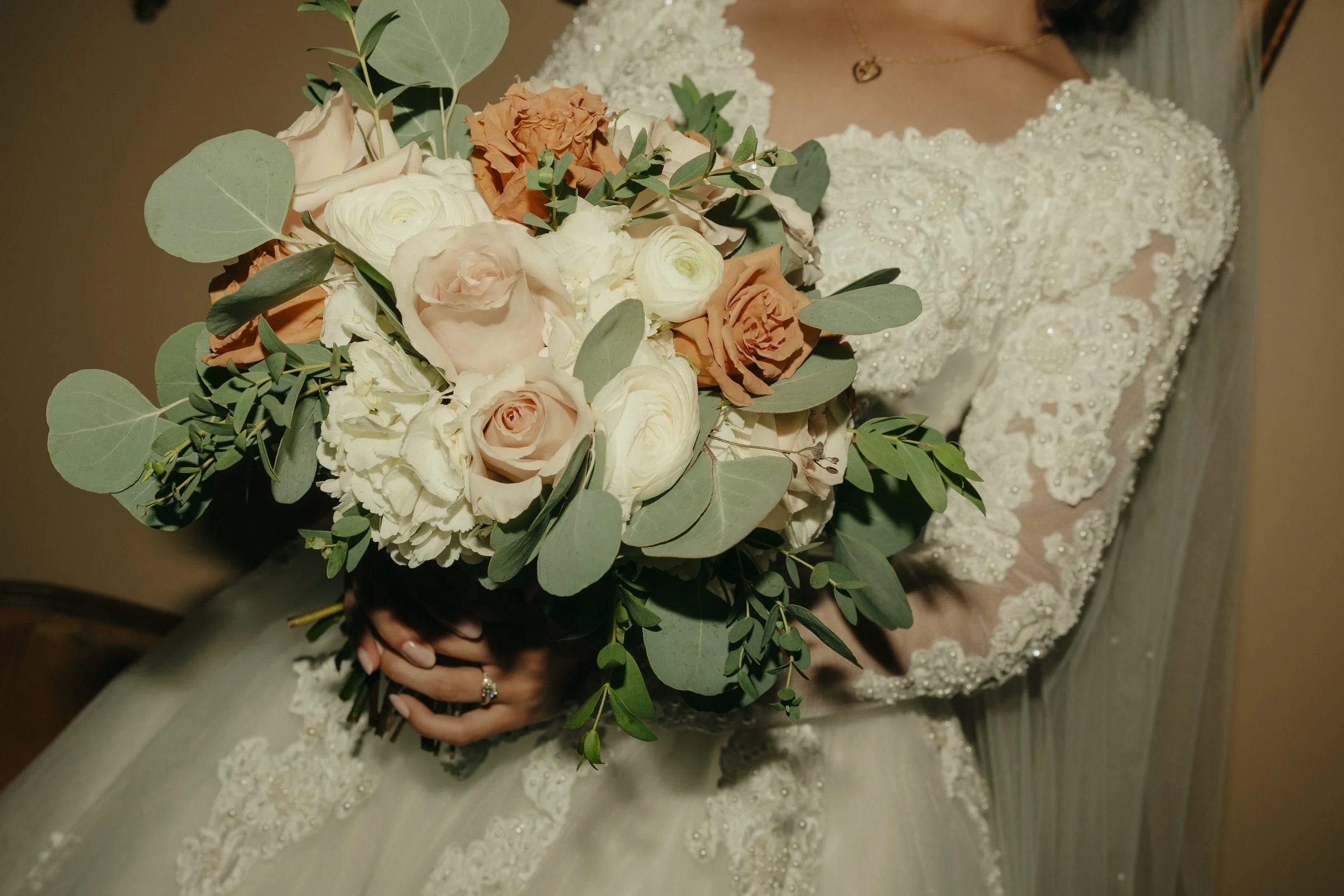 A bride holding a bouquet of pale pink, white, and peach roses, eucalyptus leaves, and other greenery. She is wearing a white wedding dress with lace and bead embellishments on the bodice.